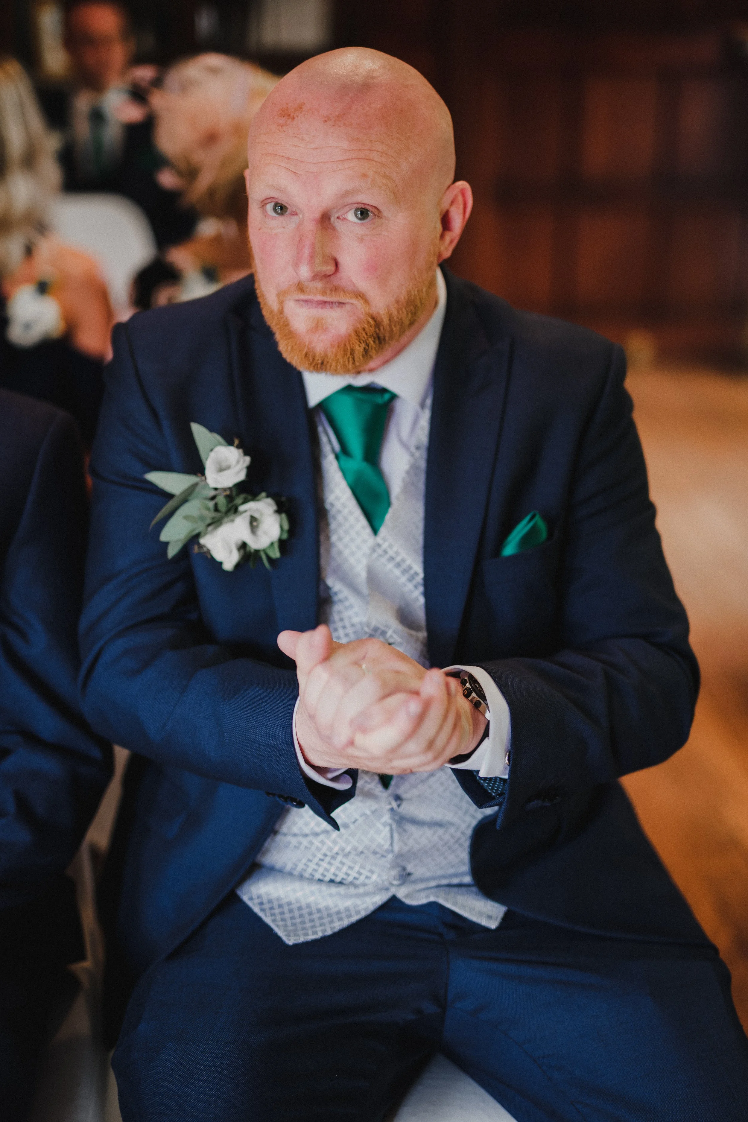 A man with a bald head and red beard wearing a navy suit, green tie, and a boutonnière, sitting at an event, with blurred people in the background.