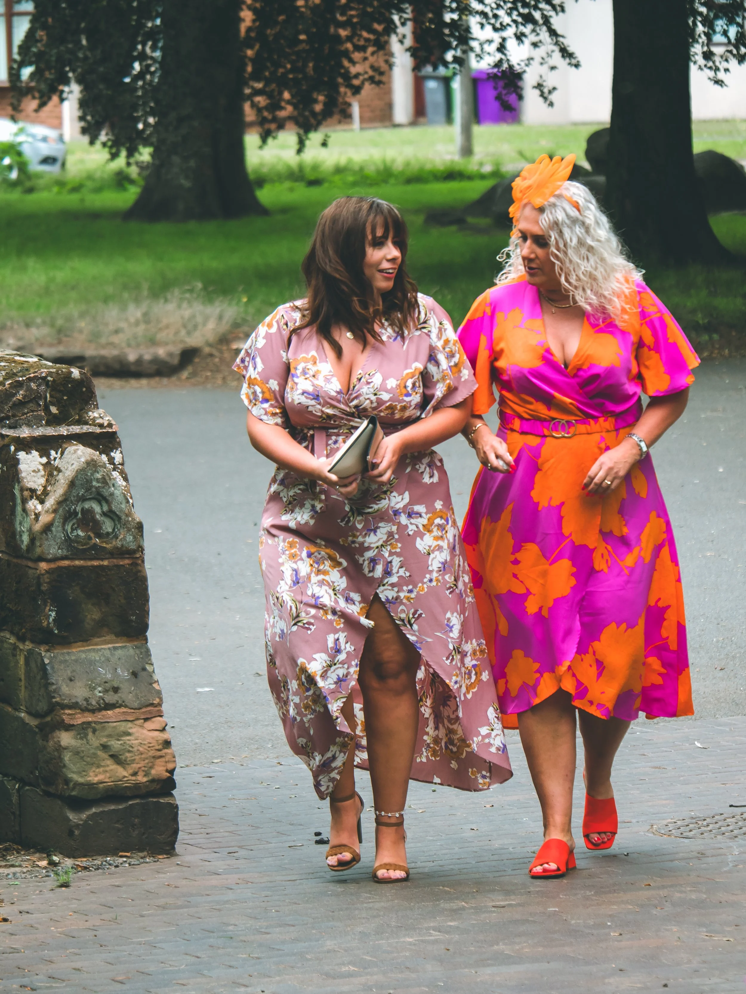 Two women walking outdoors, one in a floral pink dress and the other in a floral pink and orange dress with a hat, both wearing high heels.