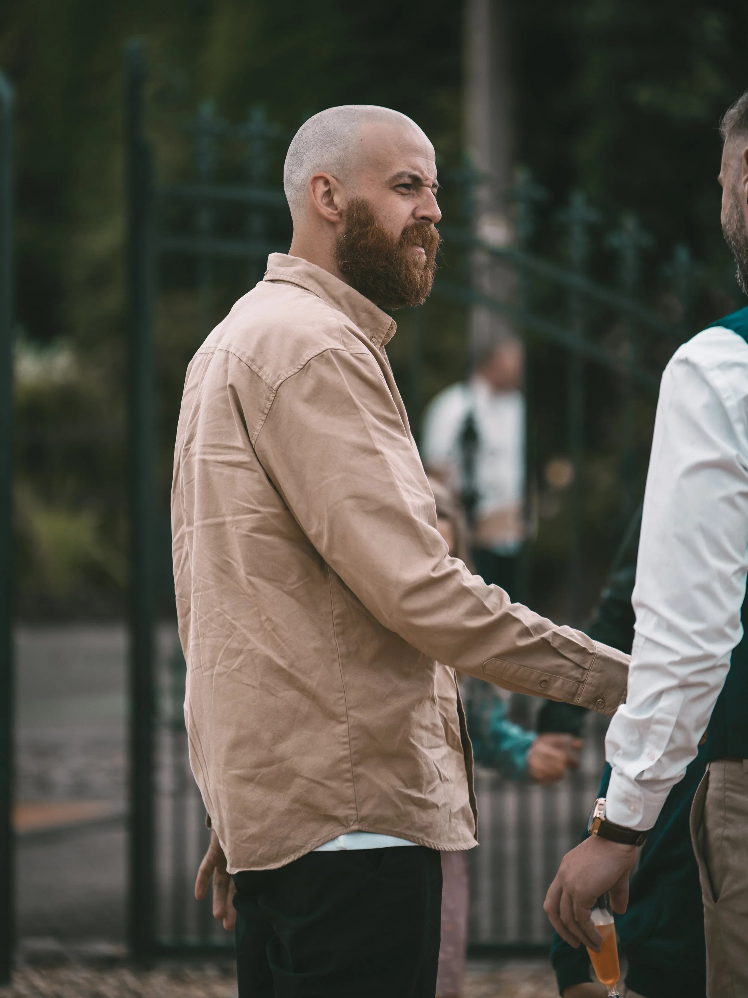 Two men holding hands and facing each other outdoors, possibly greeting or engaging in conversation, with a blurred background of trees and people.