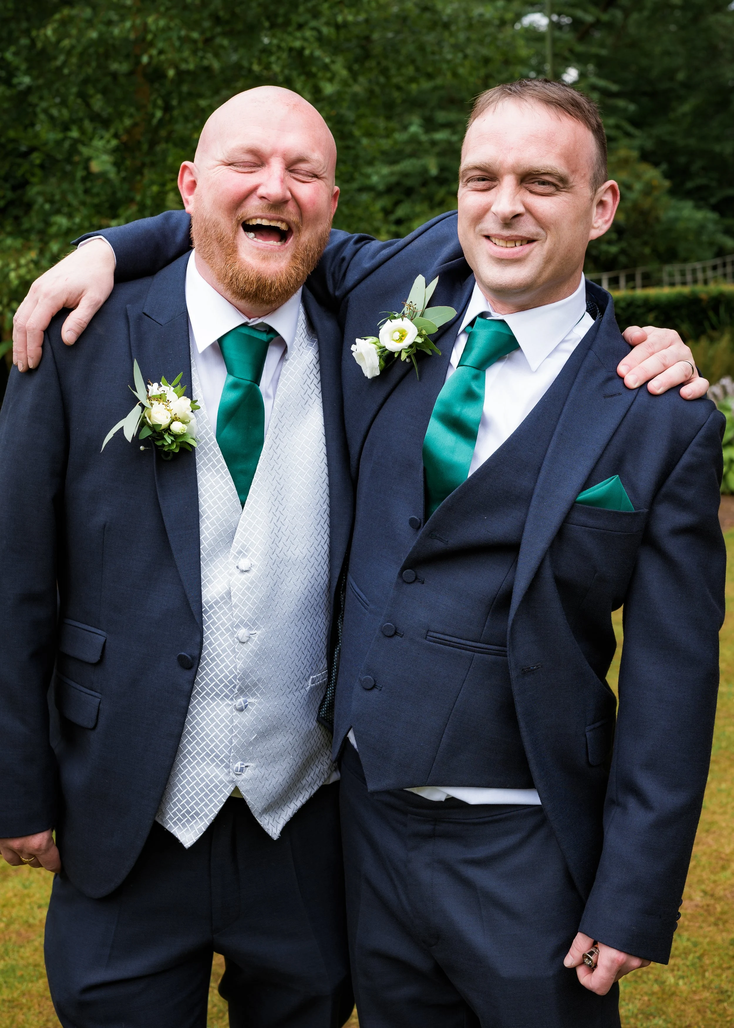 Two men in suits having fun outdoors, one with a beard and the other clean-shaven, both wearing corsages, with trees and grass in the background.