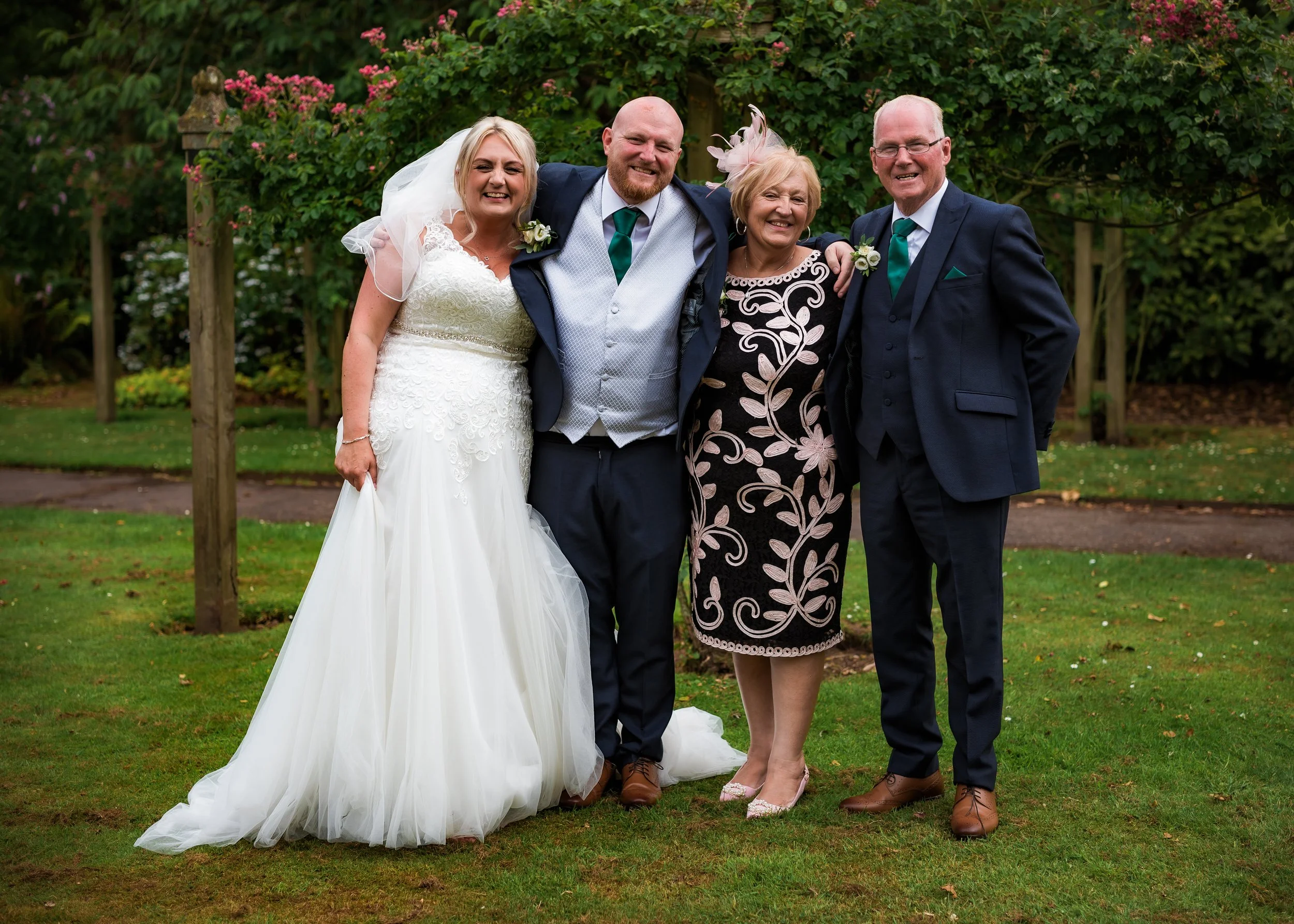 A group of five people, two women and three men, standing outdoors on grass with trees and pink flowers in the background, smiling, dressed in wedding attire.
