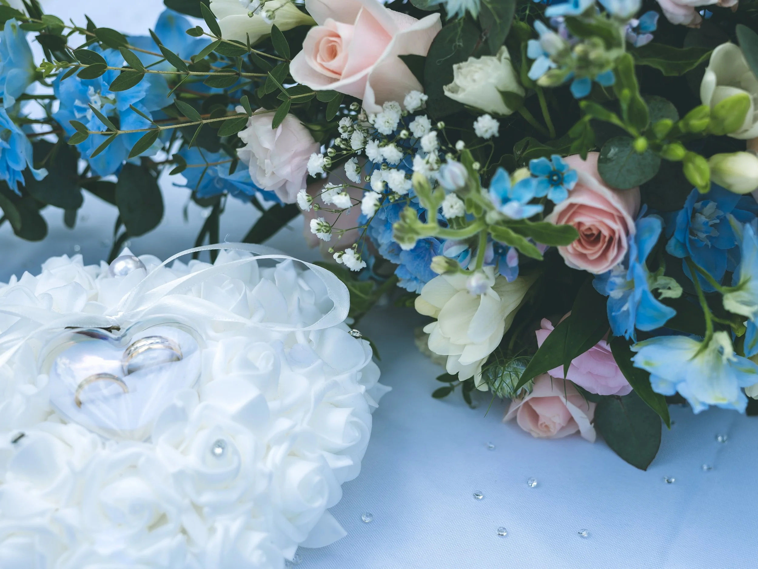 Close-up of pastel-colored roses and blue hydrangeas with greenery, white floral decorations, and small scattered rhinestones on a light blue background.