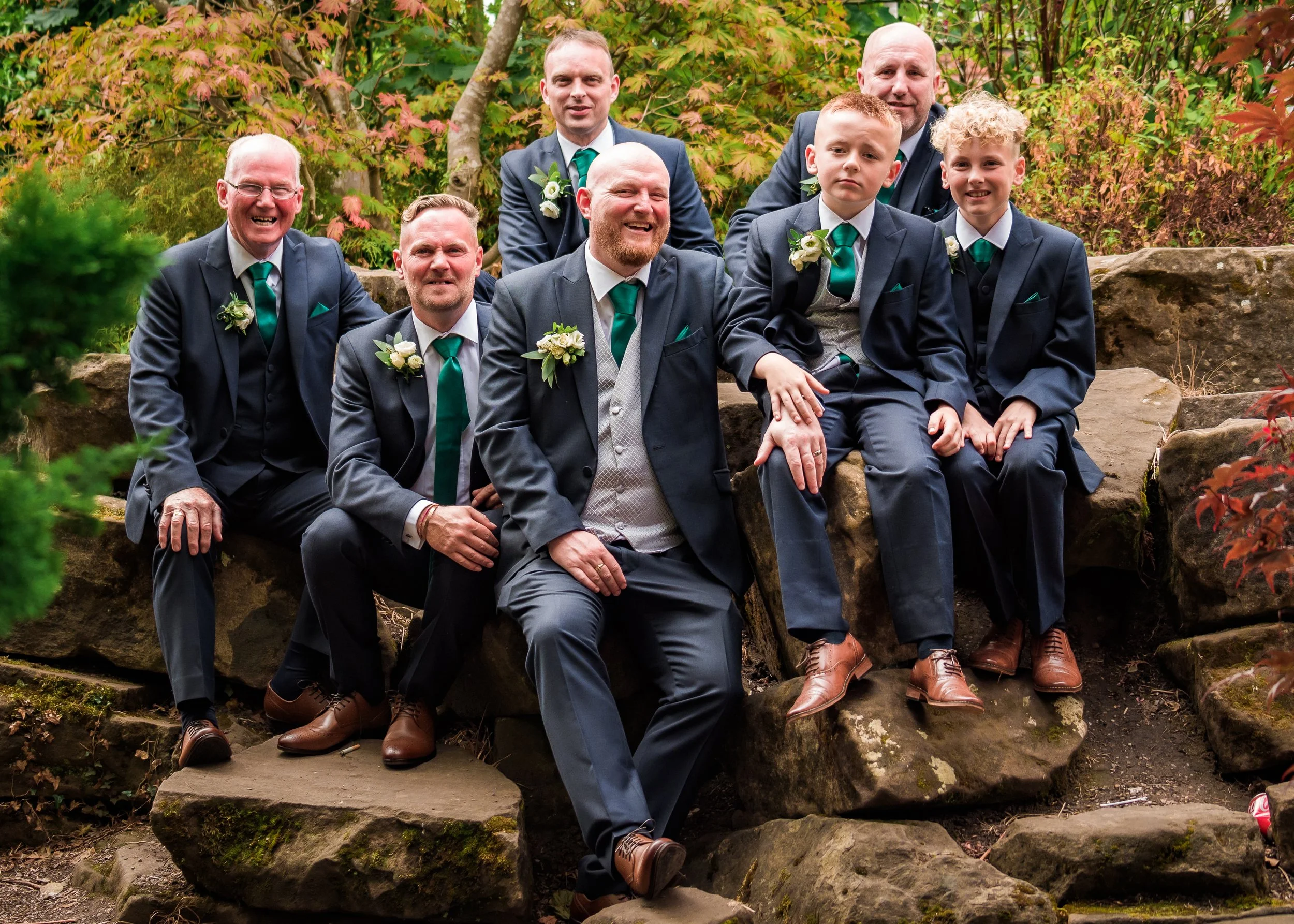 Group of men and boys in suits sitting on stone steps outdoors surrounded by trees with fall foliage