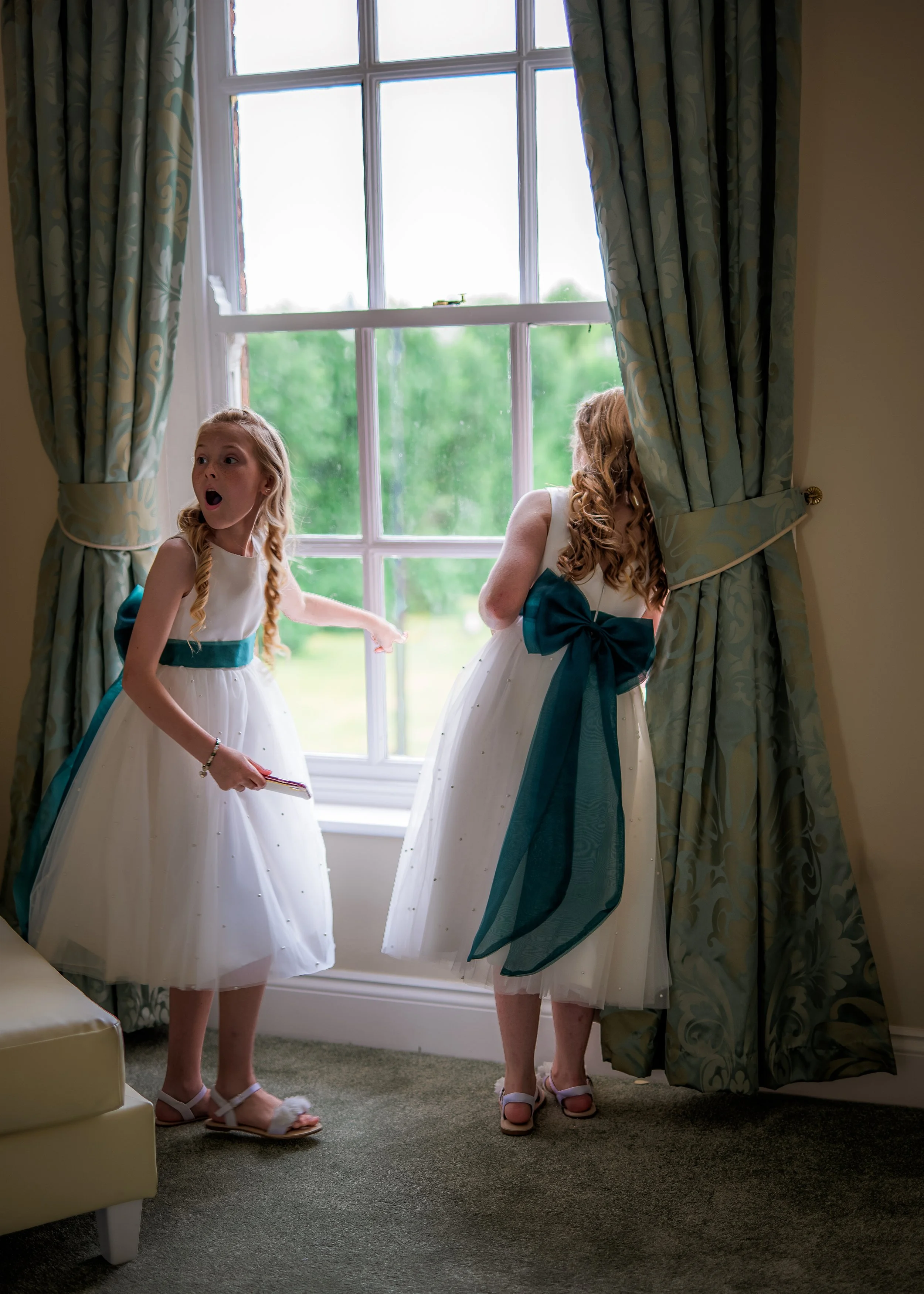 Two young girls in white dresses with teal sashes and bows, standing near a window with green scenery outside. One girl is pointing at something, and the other is leaning against the curtains.