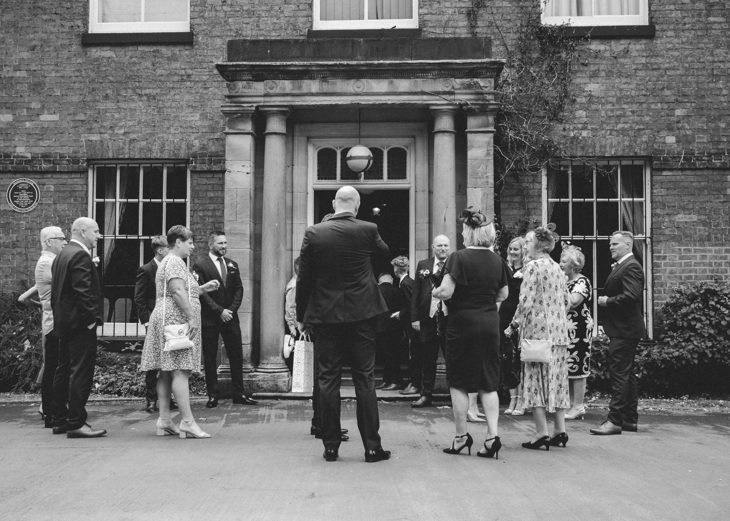 A black and white photo of a group of people outside a brick building, gathered in a semi-circle, with some dressed in formal attire, possibly attending a wedding or ceremony, and a person with a bald head and a suit standing with their back to the c