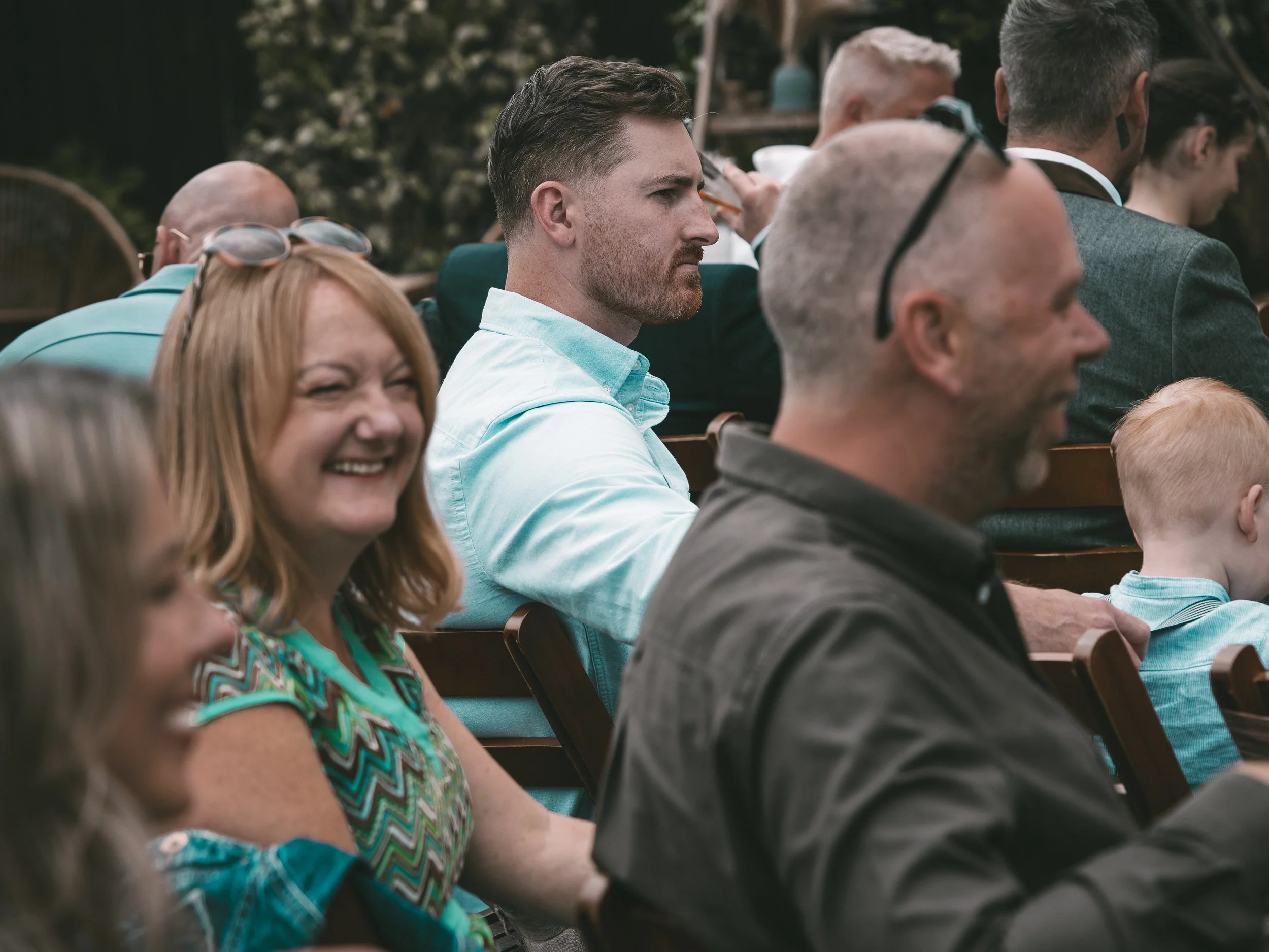 People sitting in a row outdoors, dressed in casual and semi-formal clothing, attending an event or gathering.