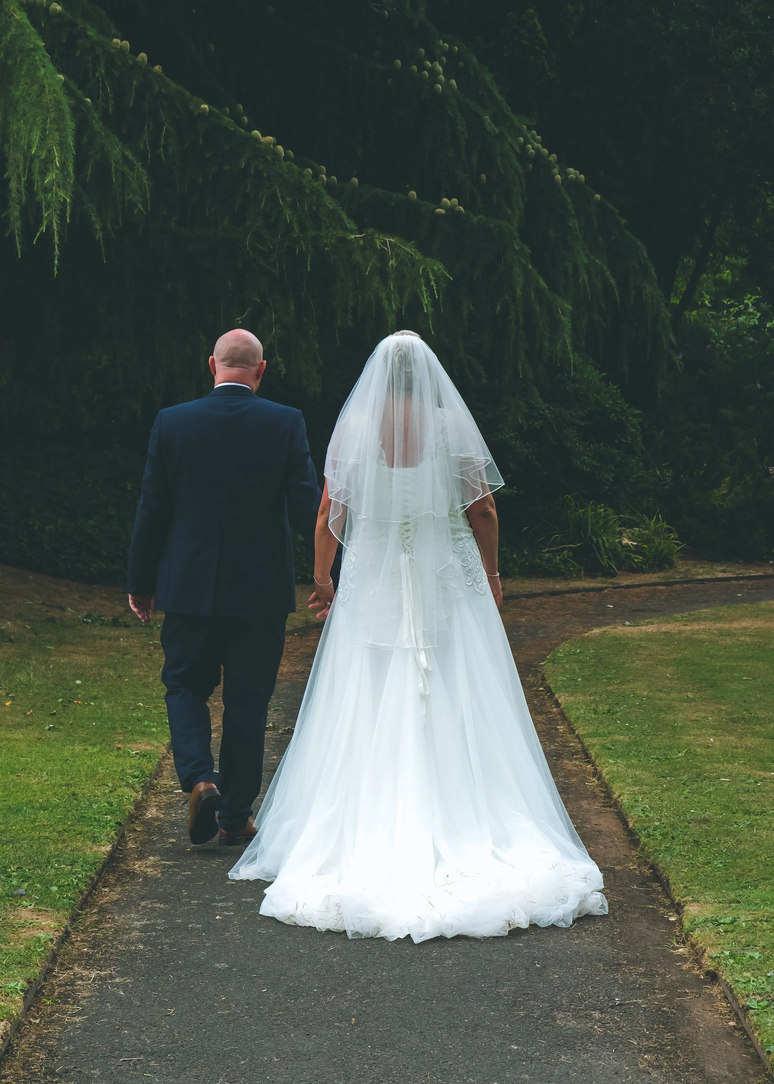 A bride and groom walking hand in hand on a pathway surrounded by greenery, with the groom in a dark suit and the bride in a white wedding gown and veil.