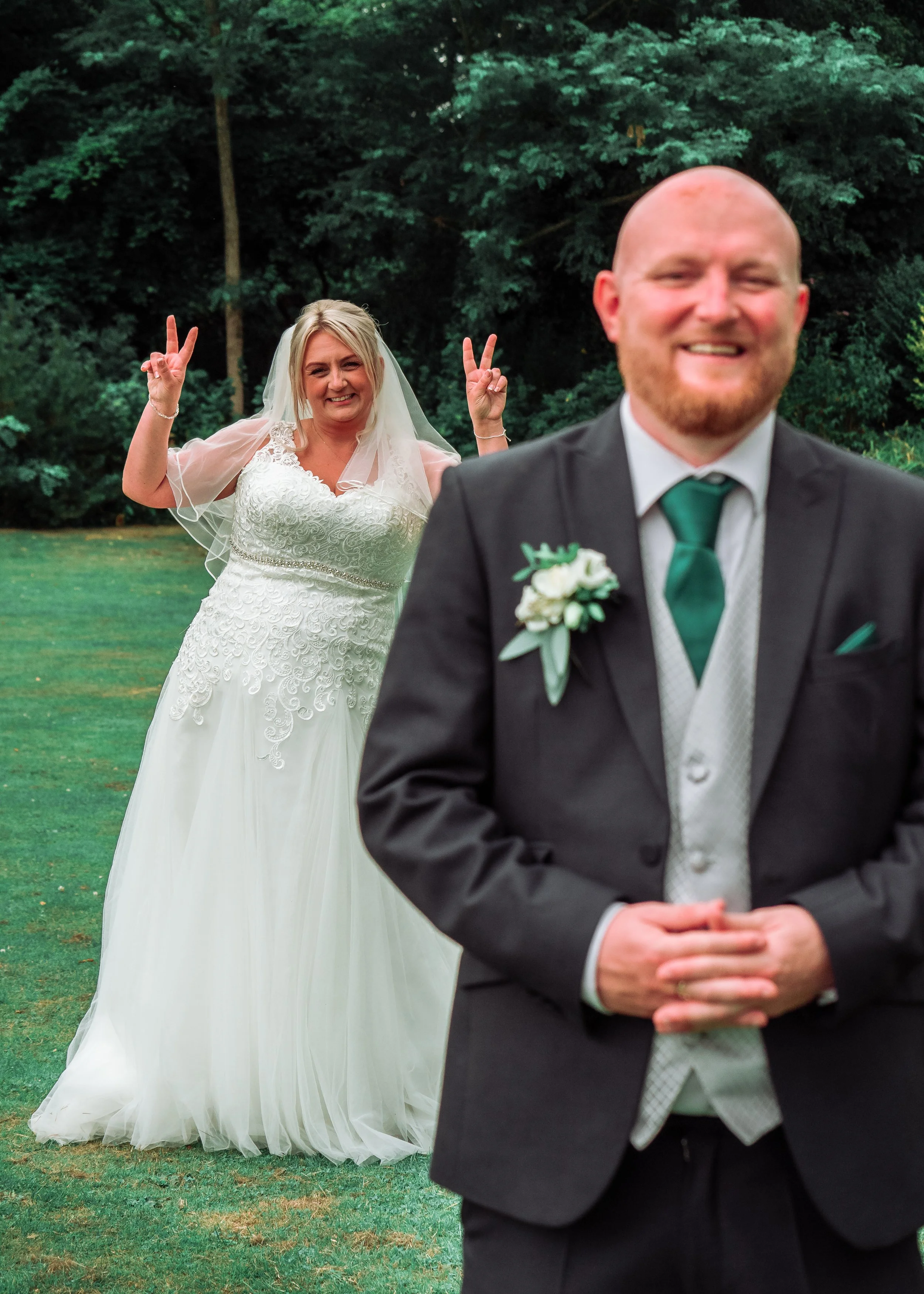 A bride making a peace sign with two fingers behind a groom at a wedding outdoors. The bride is smiling, wearing a white lace wedding dress and veil, and the groom is in a black suit with a white shirt and green tie, smiling.