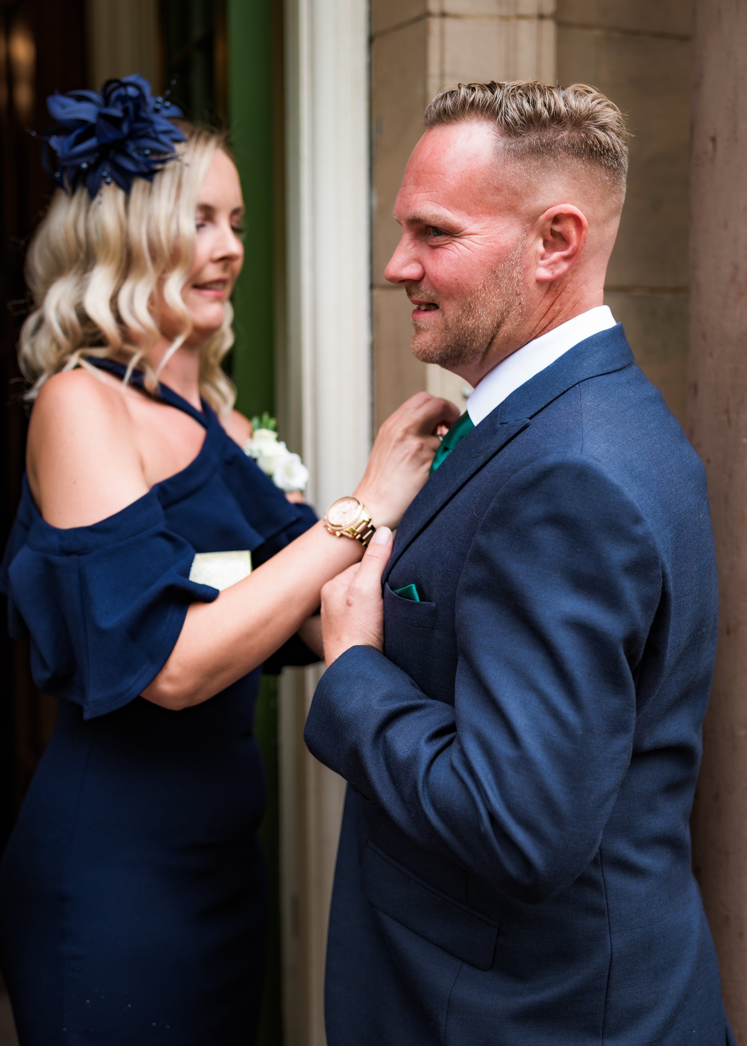 A woman in a navy dress with a large navy fabric flower on her head is adjusting the tie of a man in a blue suit. They are standing close together, smiling, in front of a stone building.