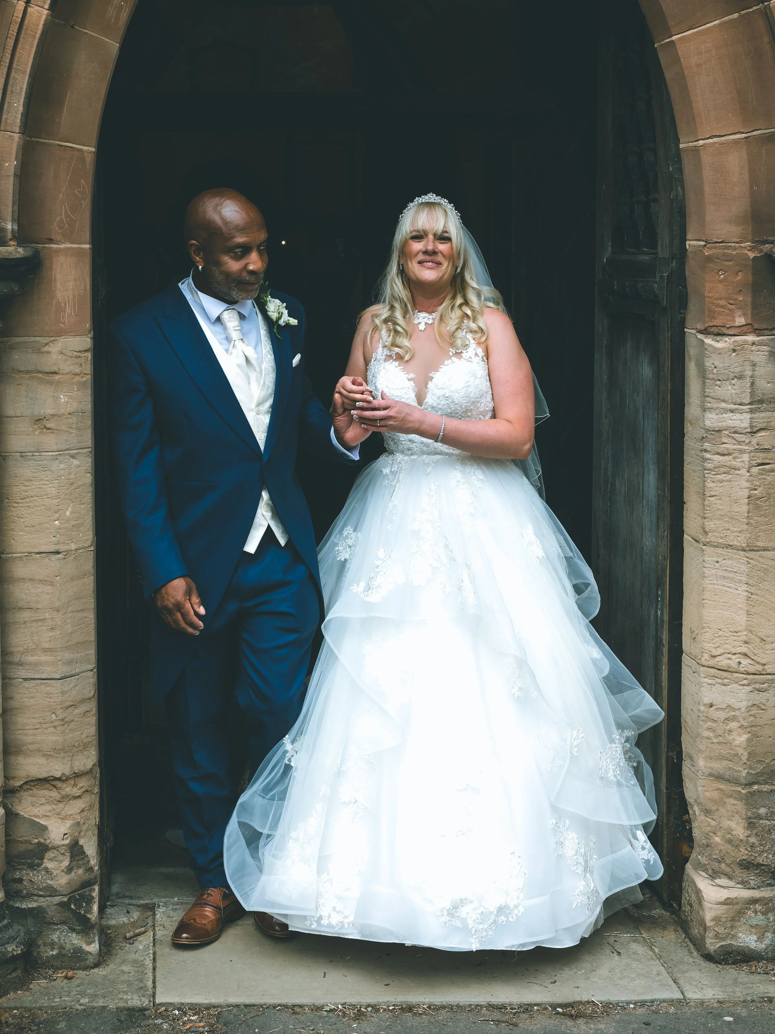 A bride and groom standing at the entrance of a church during their wedding ceremony. The bride is wearing a white lace wedding gown, a tiara, and veil, and is smiling with her hands clasped. The groom is dressed in a navy blue suit with a cream tie 