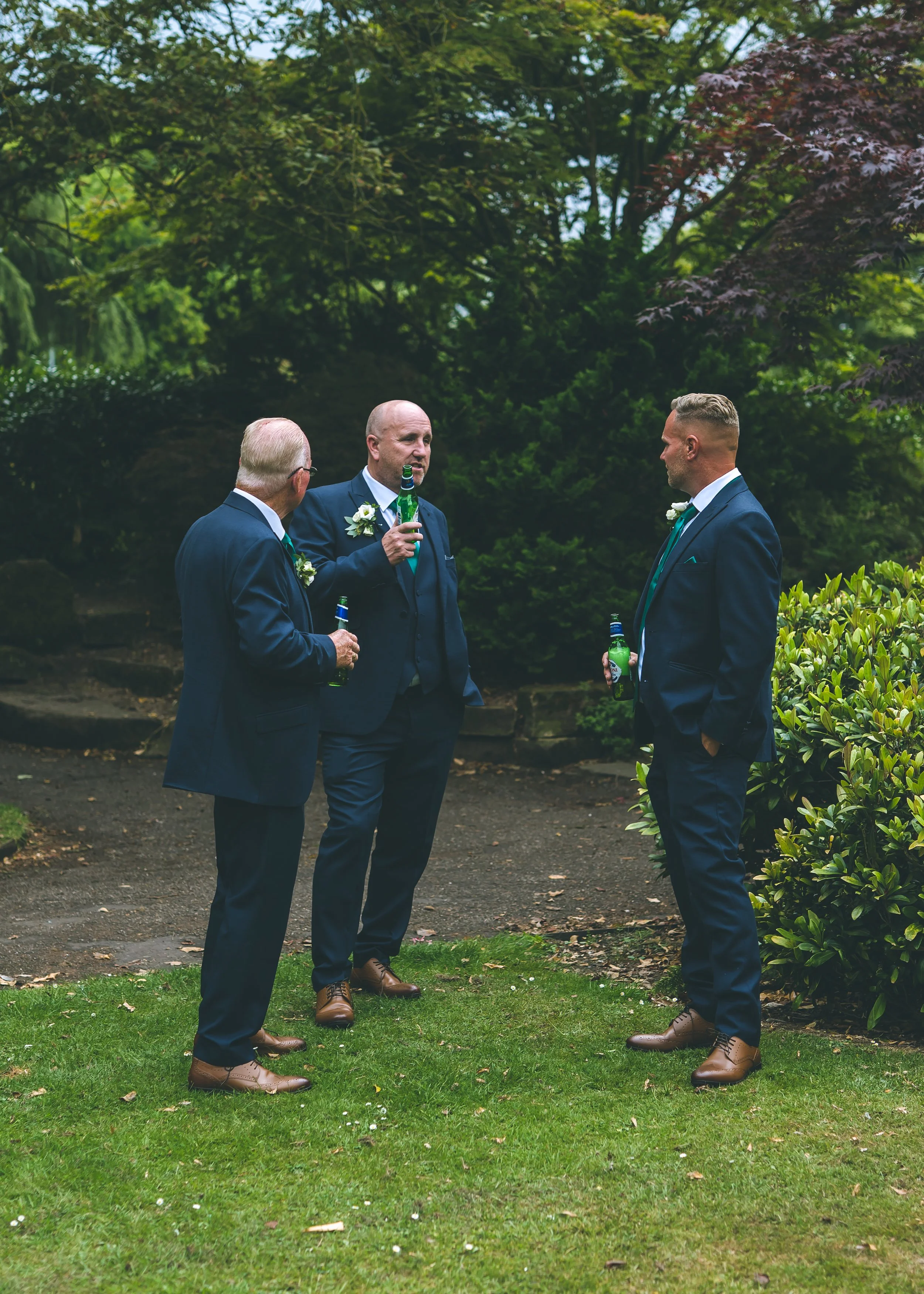 Three men in suits and brown shoes standing outdoors in a garden, holding green bottled drinks, engaged in conversation amidst green trees and bushes.