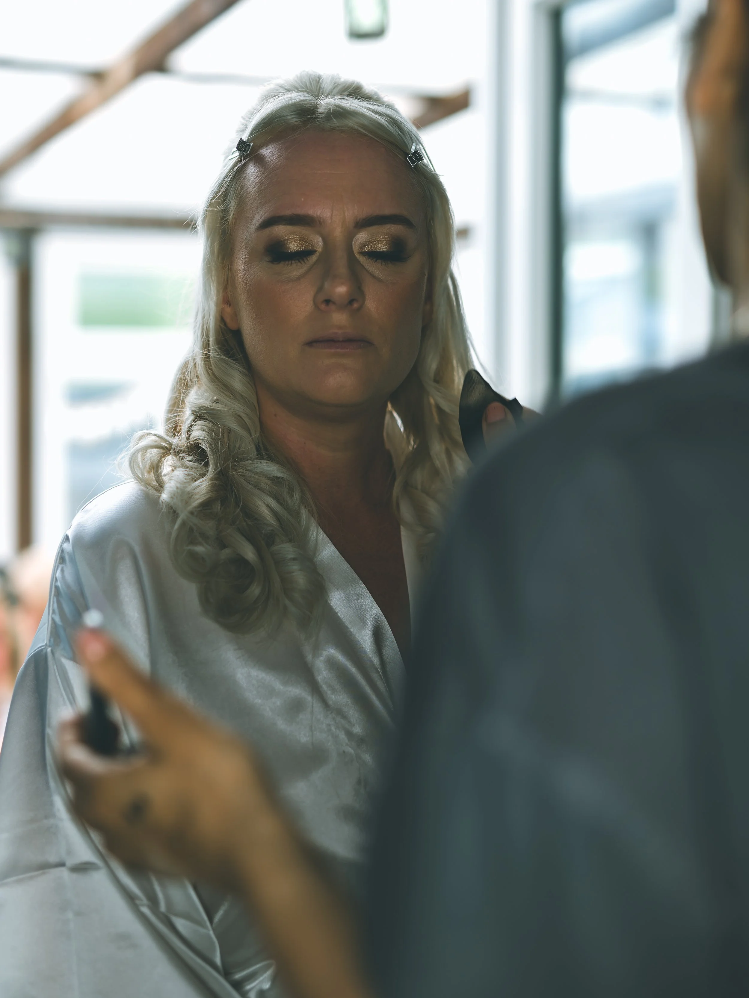 A woman with platinum blonde hair, gold eyeshadow, closed eyes, wearing a satin robe, looking into a mirror.