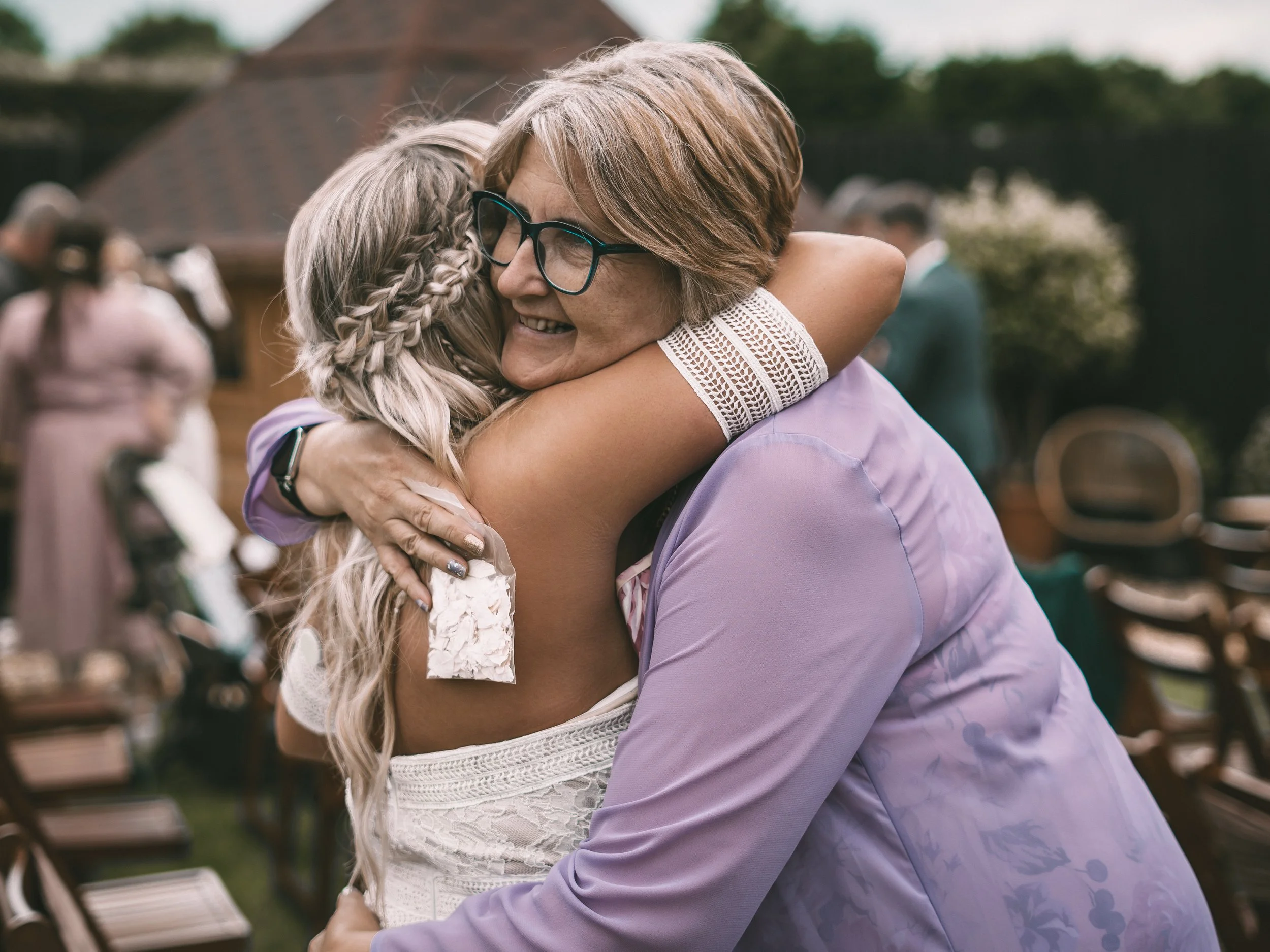 A woman with short brown hair and glasses hugging a young girl with braided blonde hair at an outdoor event.