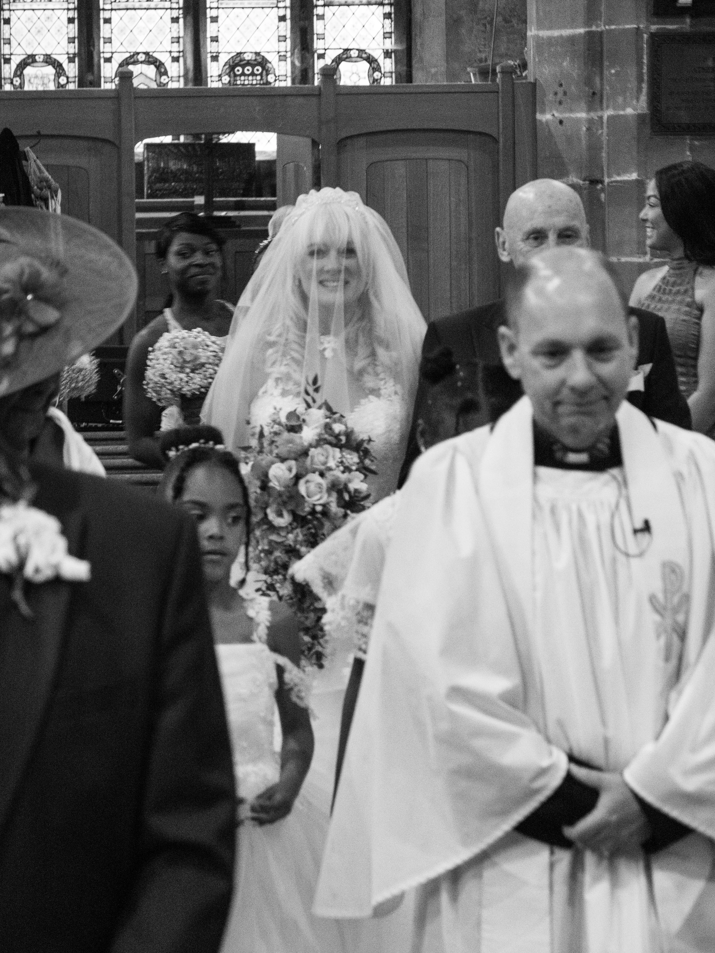Black and white photo of a group of people in a church, including a bride in a wedding dress and veil holding a bouquet, and a groom in a tuxedo. The scene appears to be during a wedding ceremony.