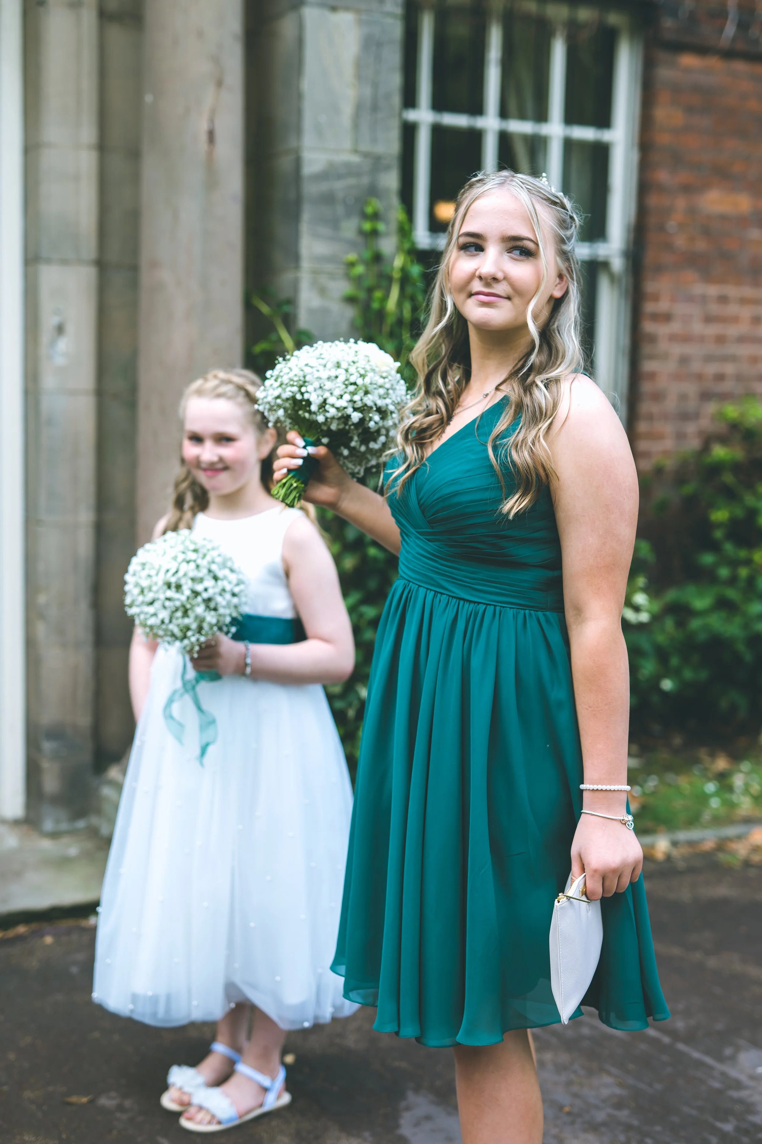 Two young women in teal dresses holding white flower bouquets outdoors in front of a brick and stone building, with trees and bushes in the background.