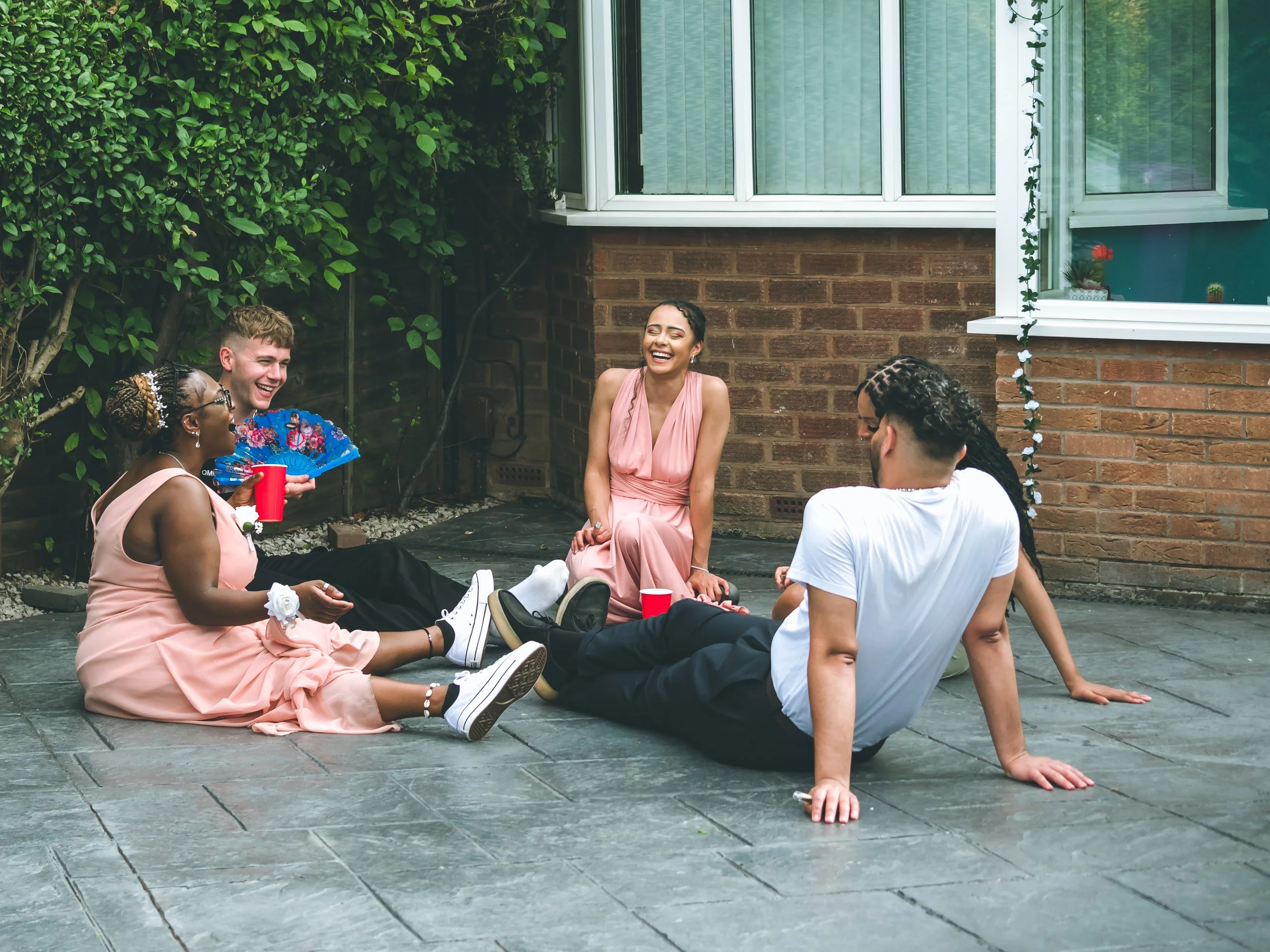 A group of five people sitting on an outdoor patio with a brick wall and window behind them, enjoying a social gathering. They are smiling and laughing, with some holding cups.