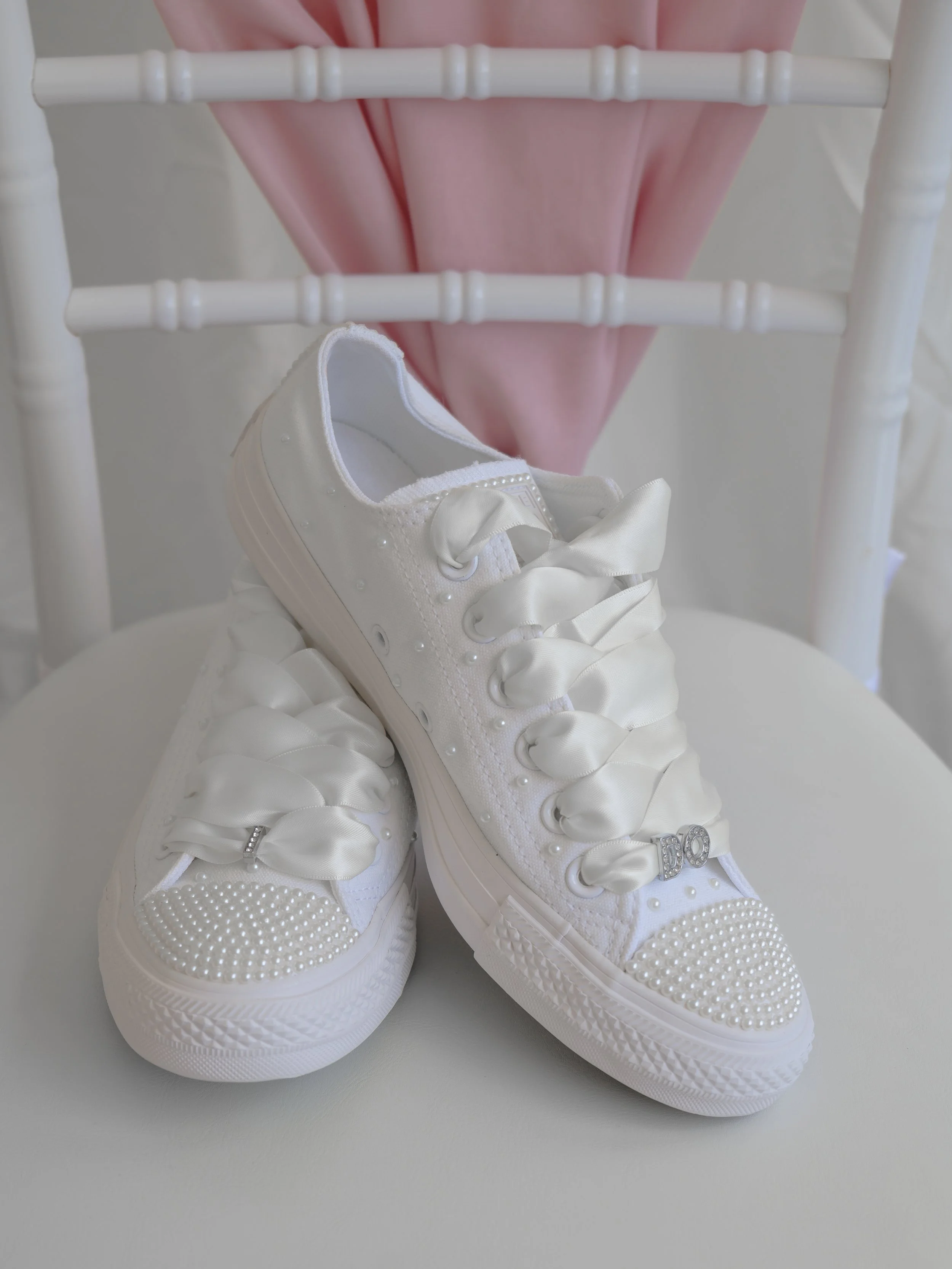 A pair of white sneakers with satin ribbons, pearl accents, and rhinestone embellishments, placed on a white surface in front of a pink fabric and a white chair.