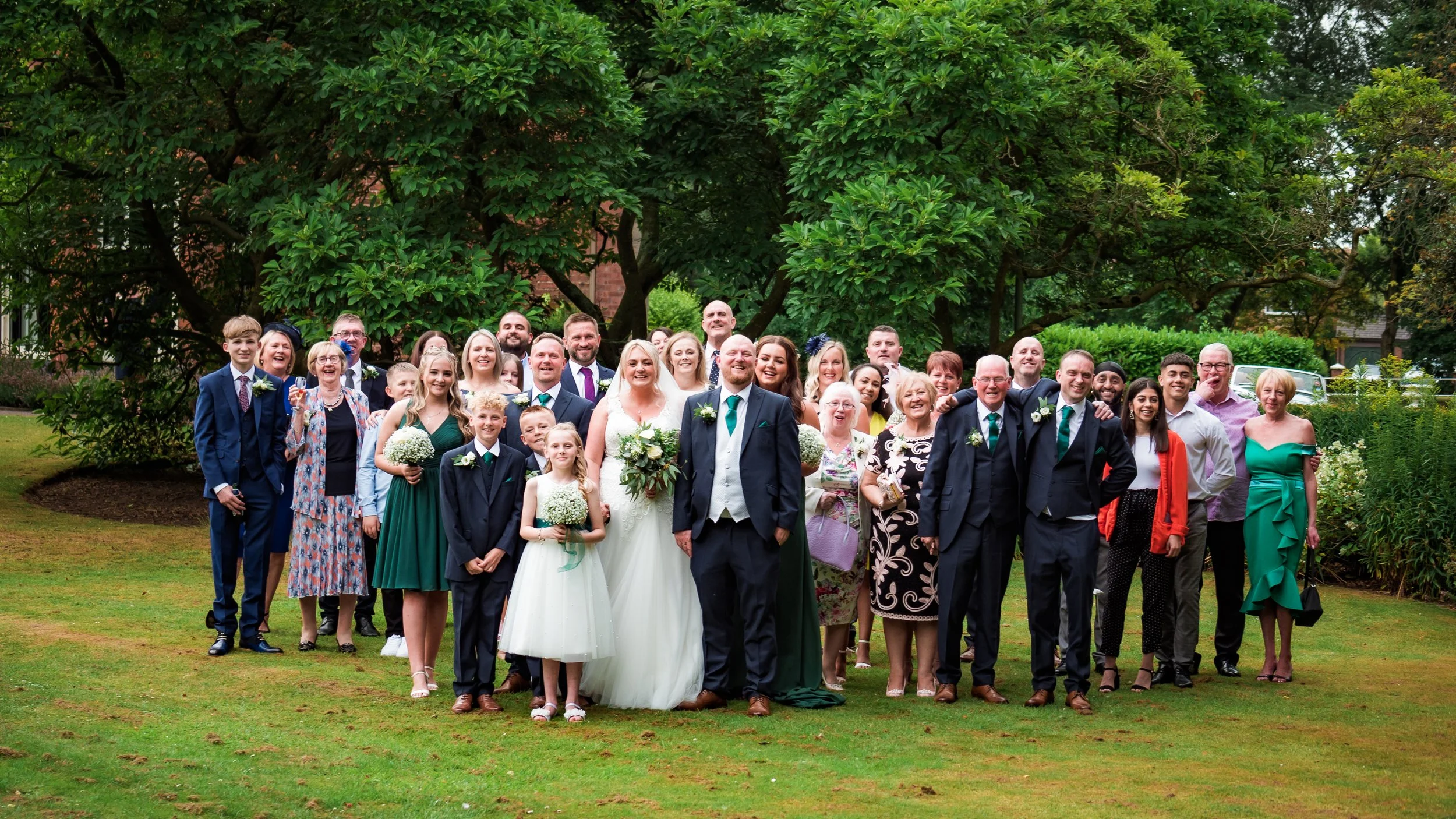 A large group of people gathered outdoors in front of a green tree, celebrating a wedding. The bride and groom are in the center, smiling, with the bride in a white dress holding a bouquet. The group includes men, women, children, and elderly, dresse