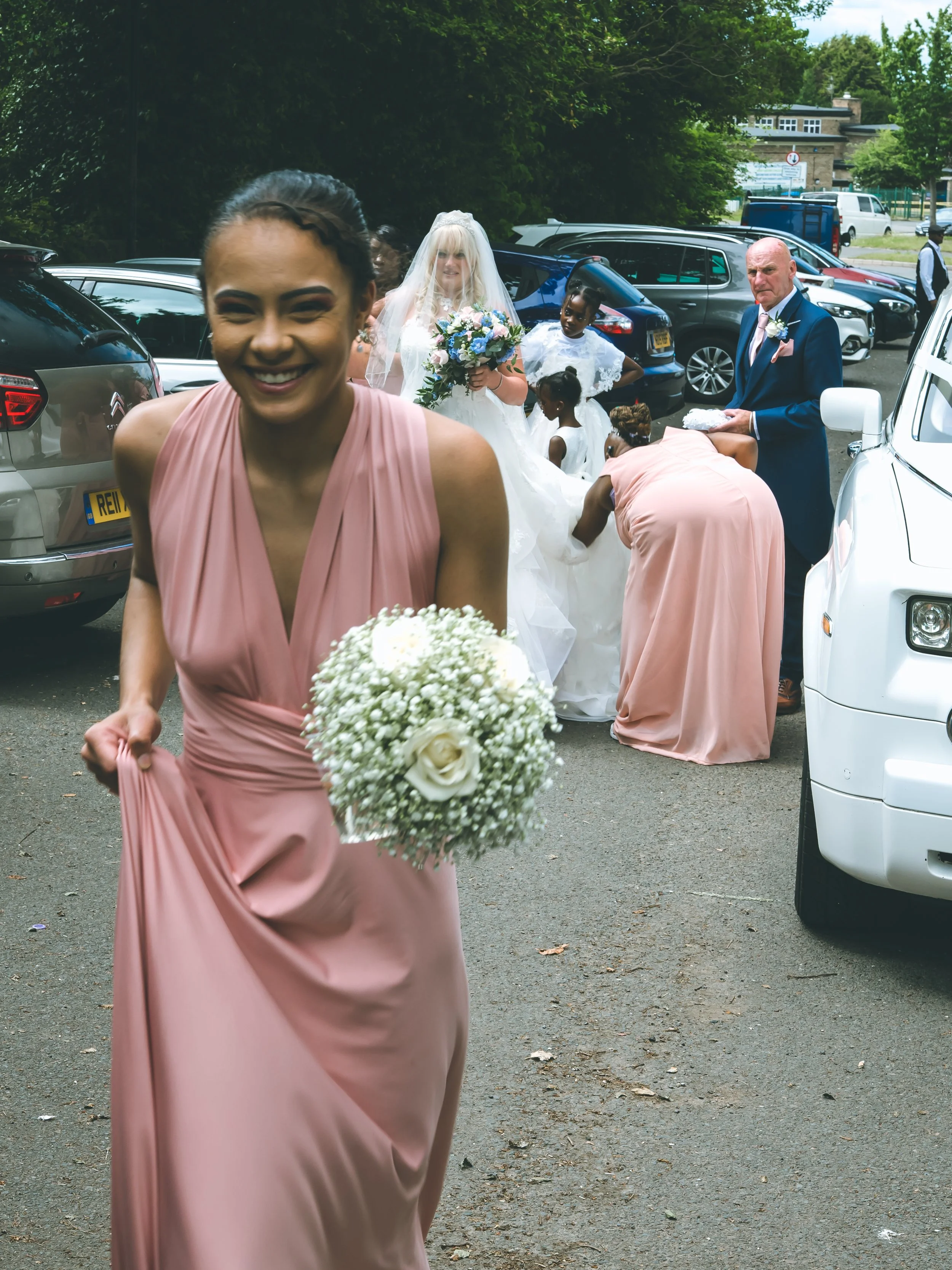 A smiling woman in a pink dress holding a bouquet of white flowers, walking in a parking lot with a wedding group in the background; a bride in a wedding dress with a veil holding a bouquet, and an older man in a suit and pink tie among others.