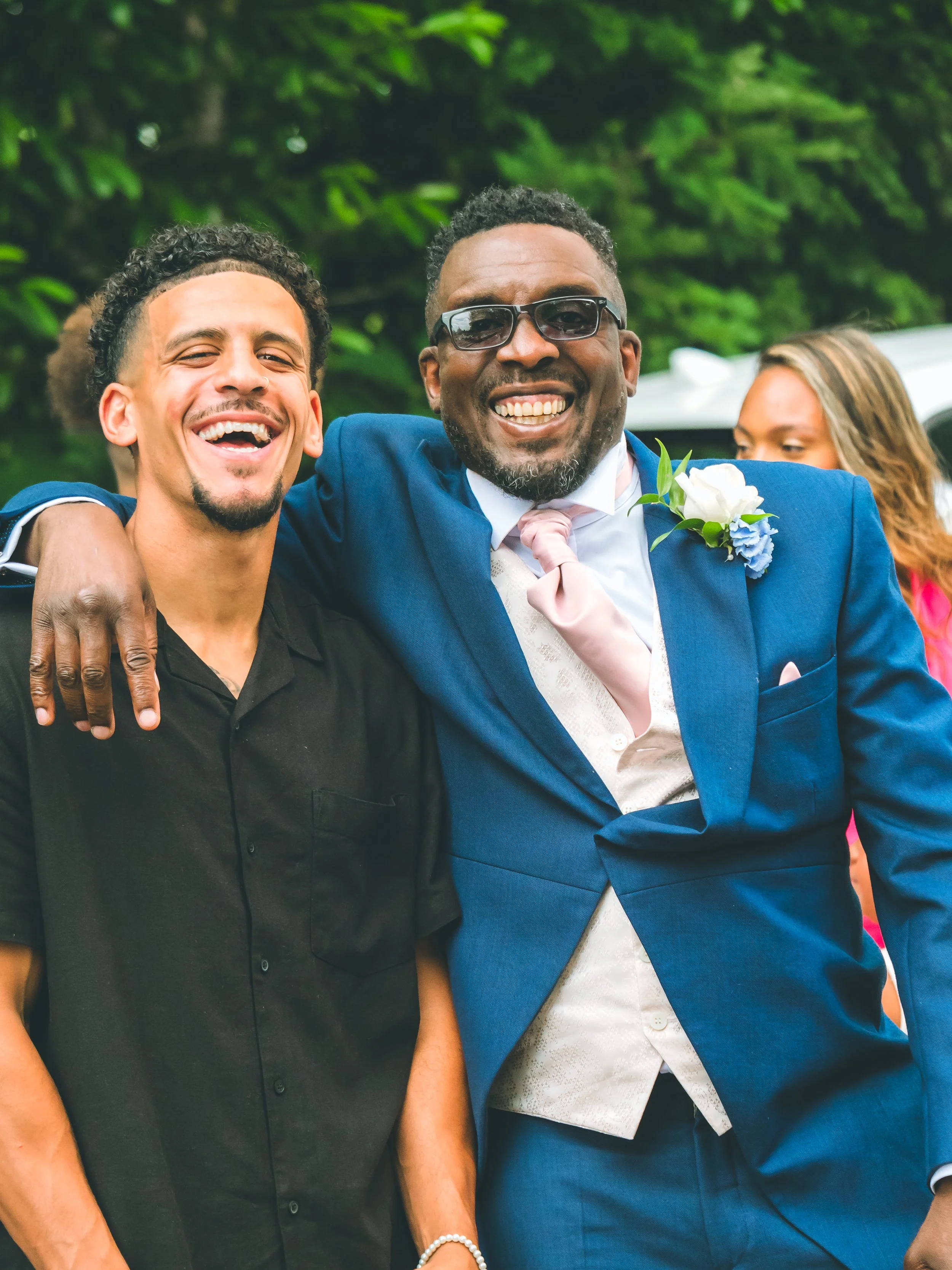 Two men standing closely together outdoors, smiling and celebrating. One man has dark skin, glasses, gray beard, wearing a blue suit with a white shirt and pink tie, with a white flower boutonniere. The other man has light skin, short curly dark hair
