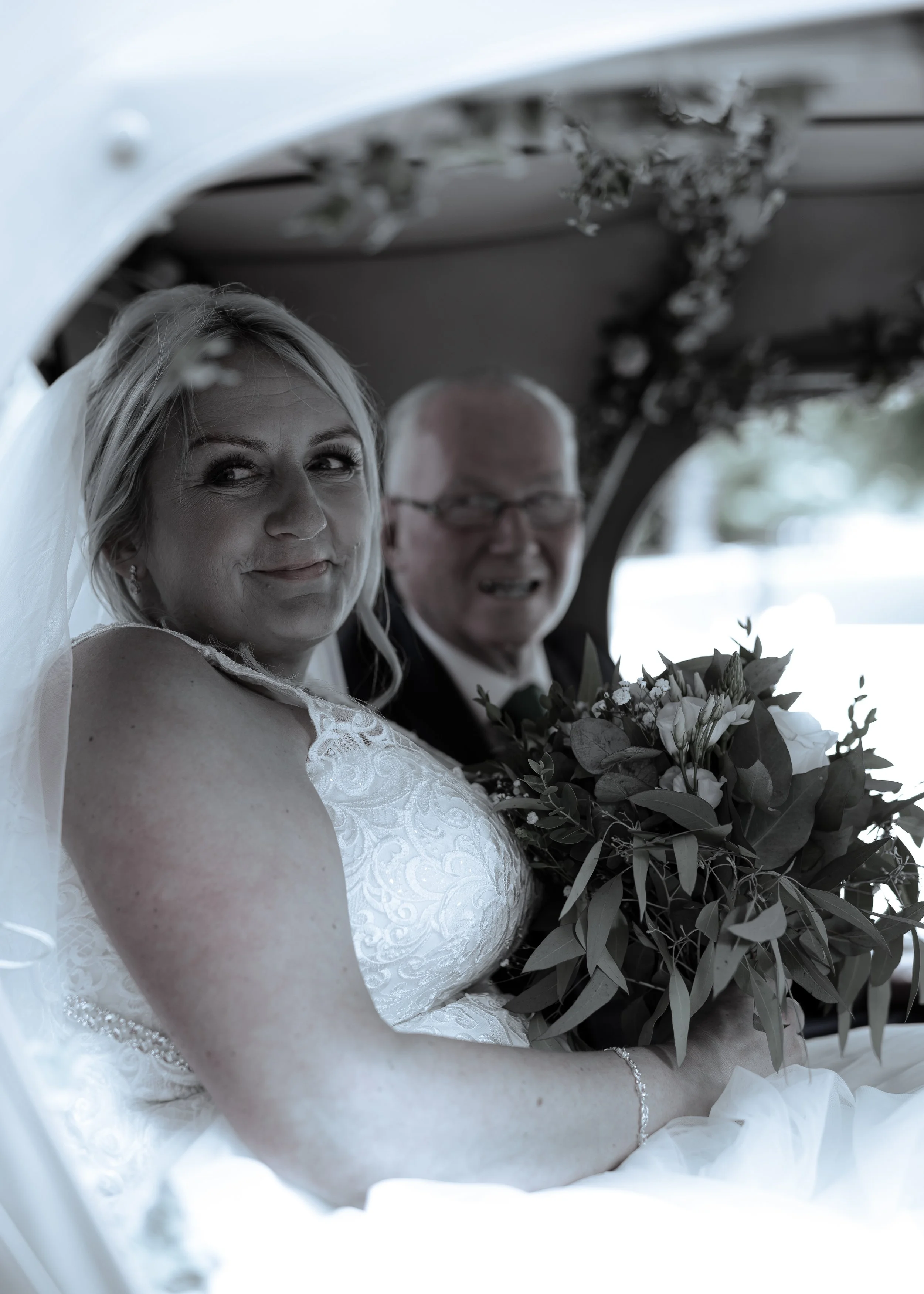 A bride and groom sit inside a car, with the bride smiling and holding a bouquet of flowers. The bride wears a white lace wedding dress, and the groom wears a tuxedo with glasses. The photo is in black and white.