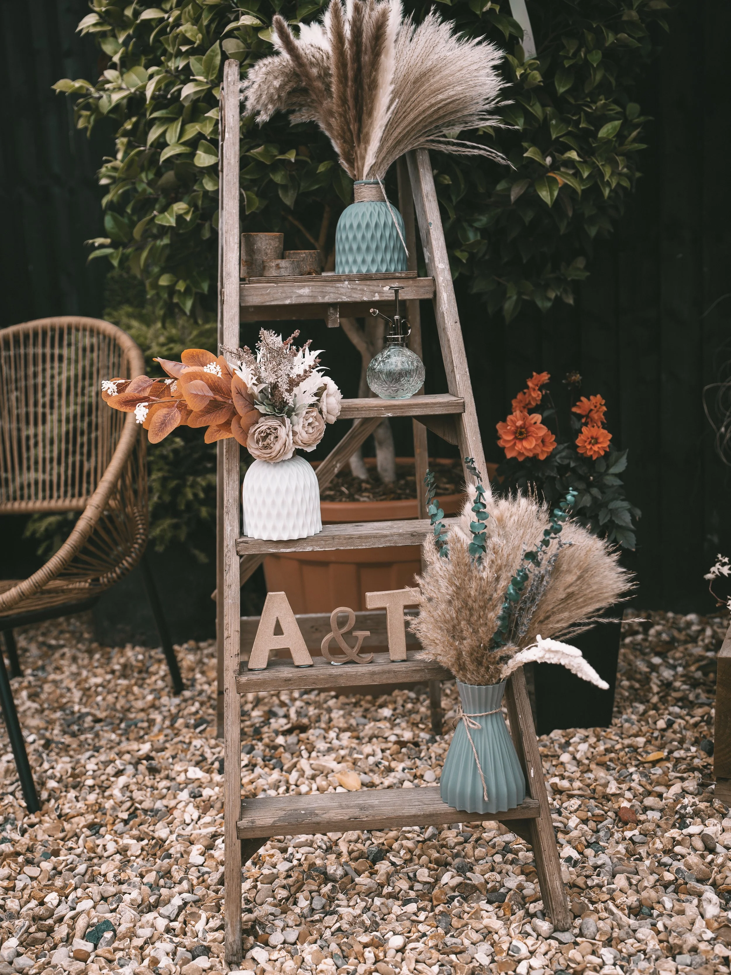 Decorative outdoor scene with a rustic wooden ladder holding vases with flowers, a glass spray bottle, and decorative letters 'A & T'. Potted plants and flowers around, gravel ground.