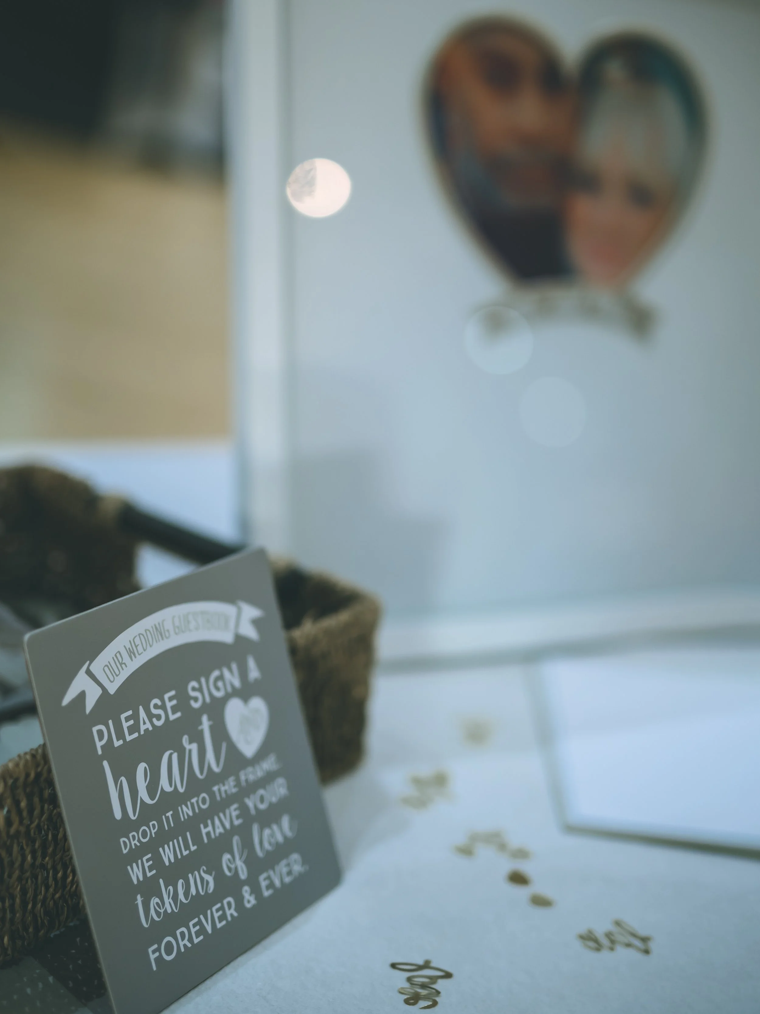 Close-up of a wedding or love-themed sign on a table, with a blurred background featuring a heart-shaped object with photos inside and a visible moon in the sky.