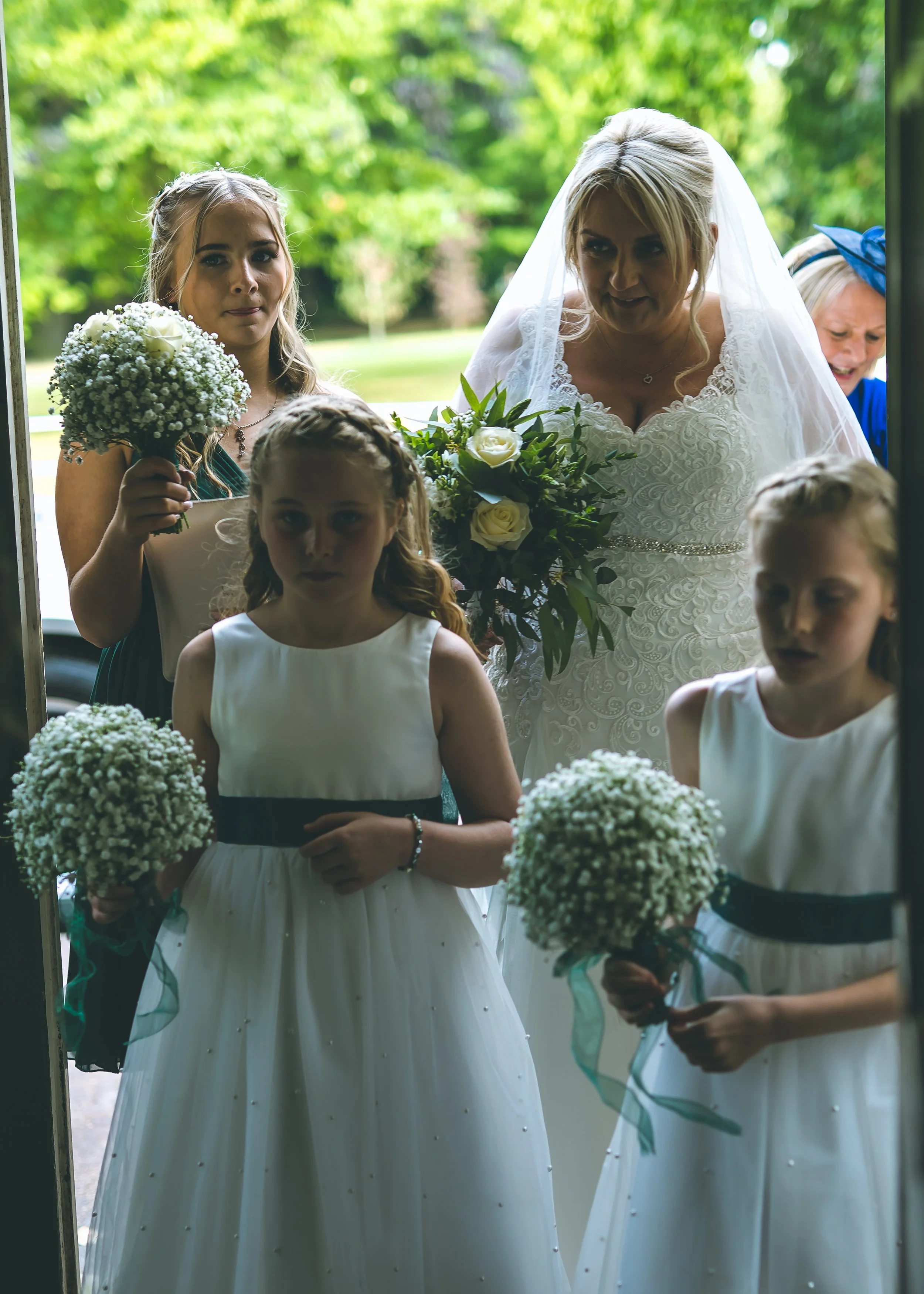 A bride in a white lace wedding dress with a veil holding a bouquet of white roses and greenery standing behind a glass door with three flower girls in white dresses with black sashes holding small bouquets of baby's breath, outdoors on a sunny day w