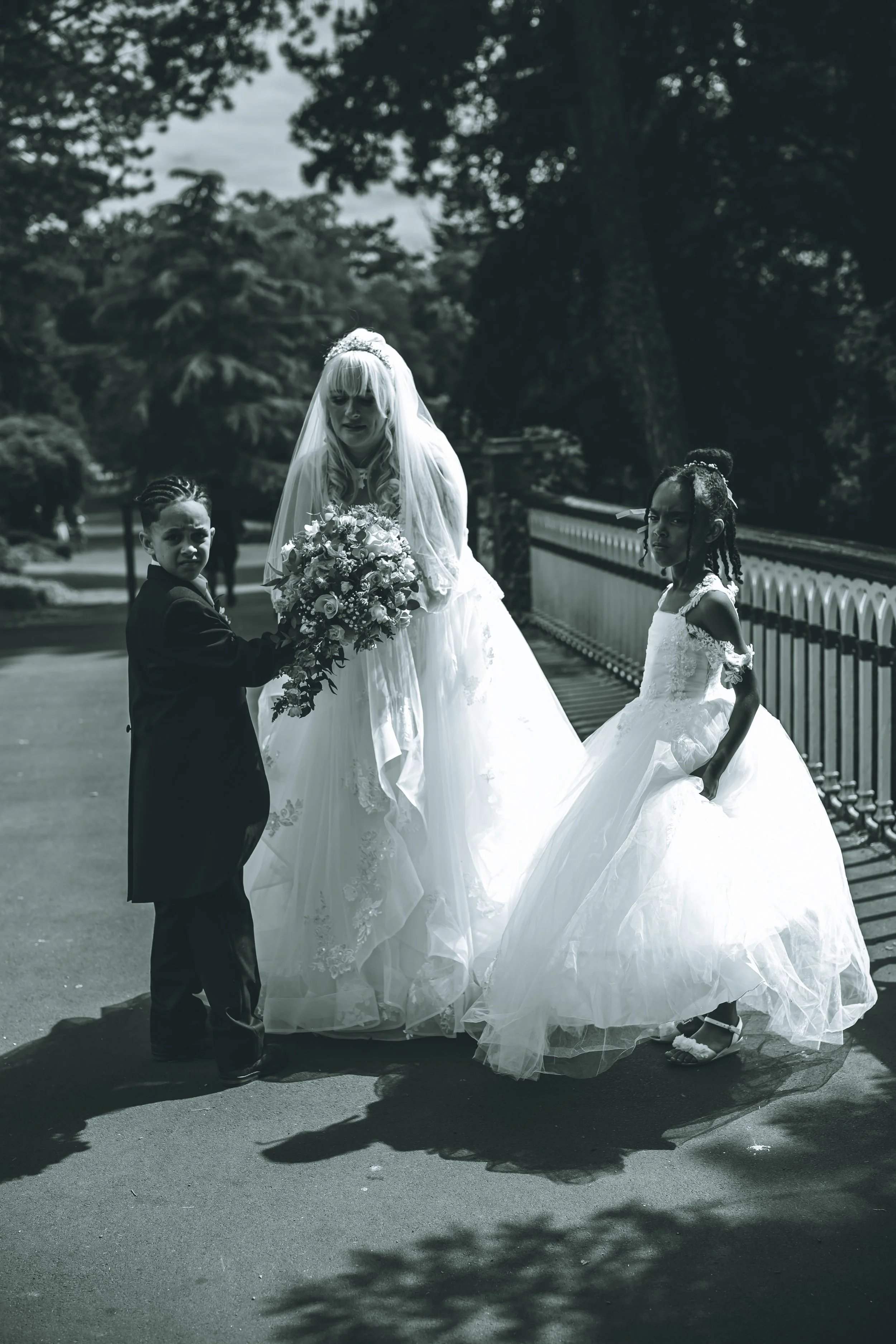 A bride in a wedding dress with two young children, a boy and a girl, outdoors on a cloudy day. The boy in a tuxedo holds a bouquet of flowers. The girl in a white dress stands beside the bride near a white fence, with trees in the background.