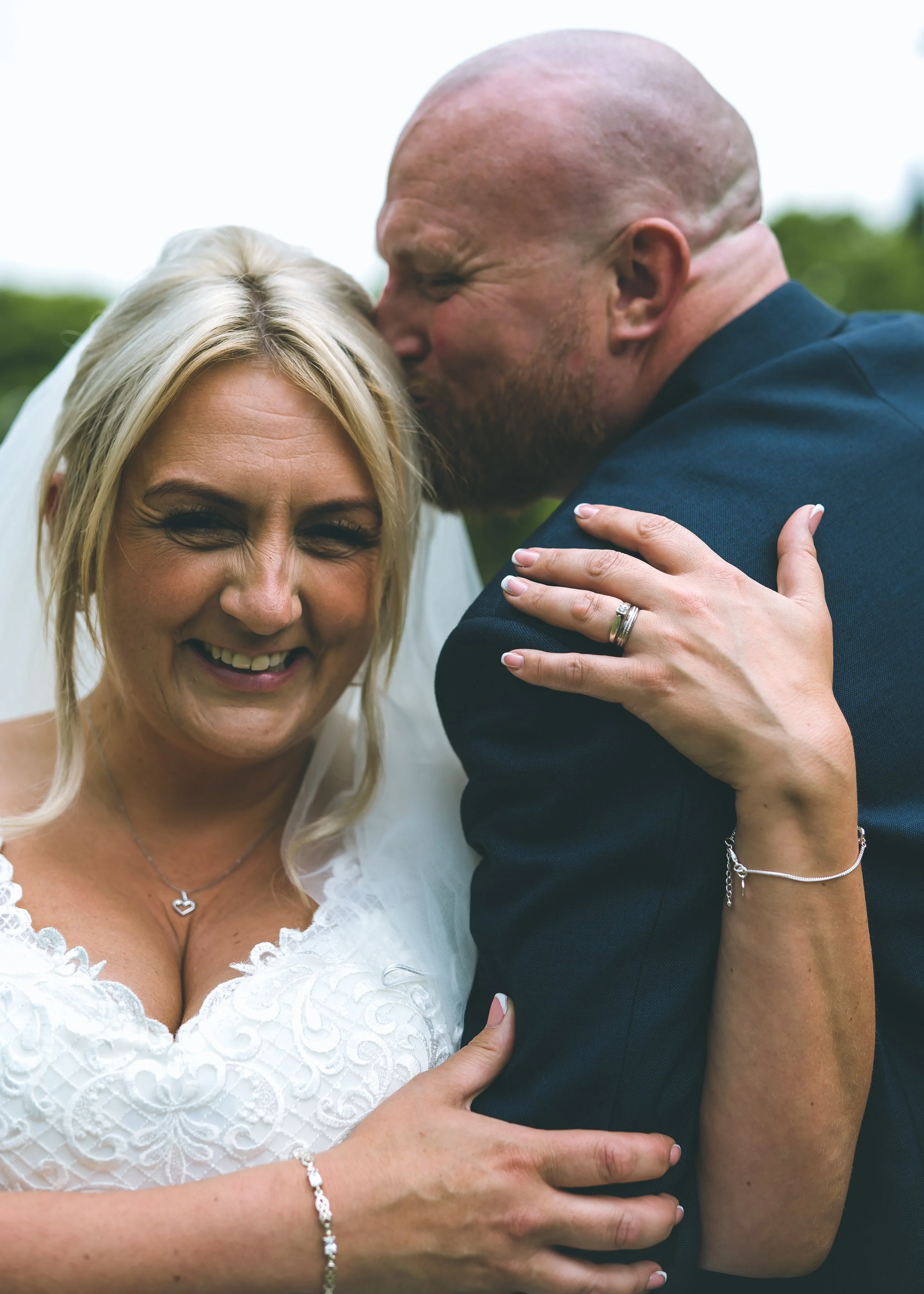 A man and woman share a loving embrace outdoors, with the man kissing the woman's forehead, both smiling and with blonde hair.