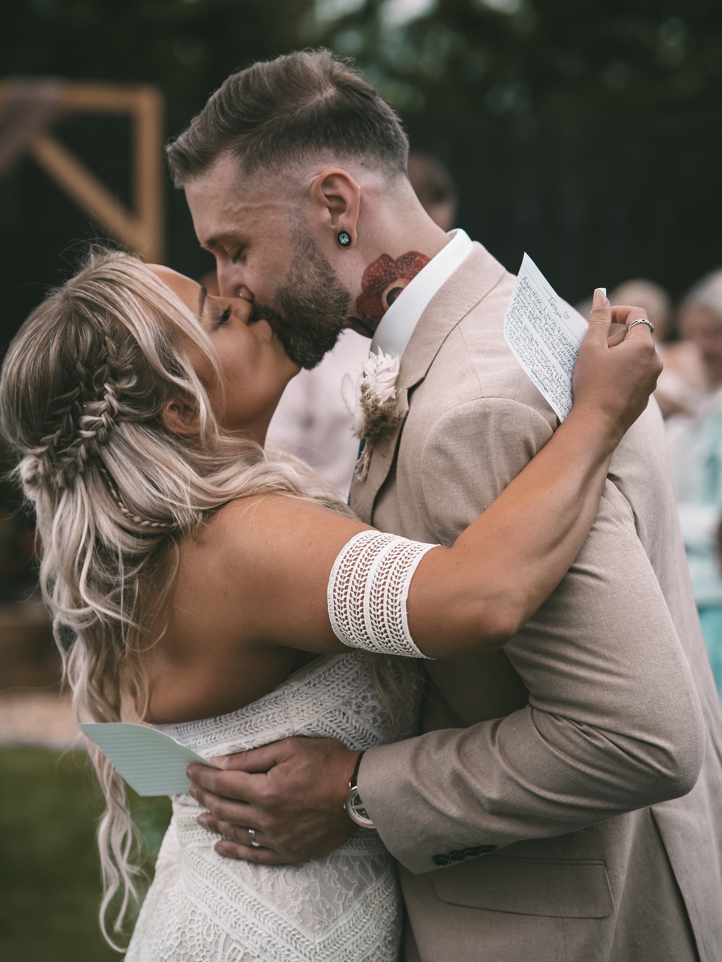 A couple sharing a kiss during their wedding ceremony outdoors, with the bride holding a handwritten note and the groom embracing her in a tan suit.