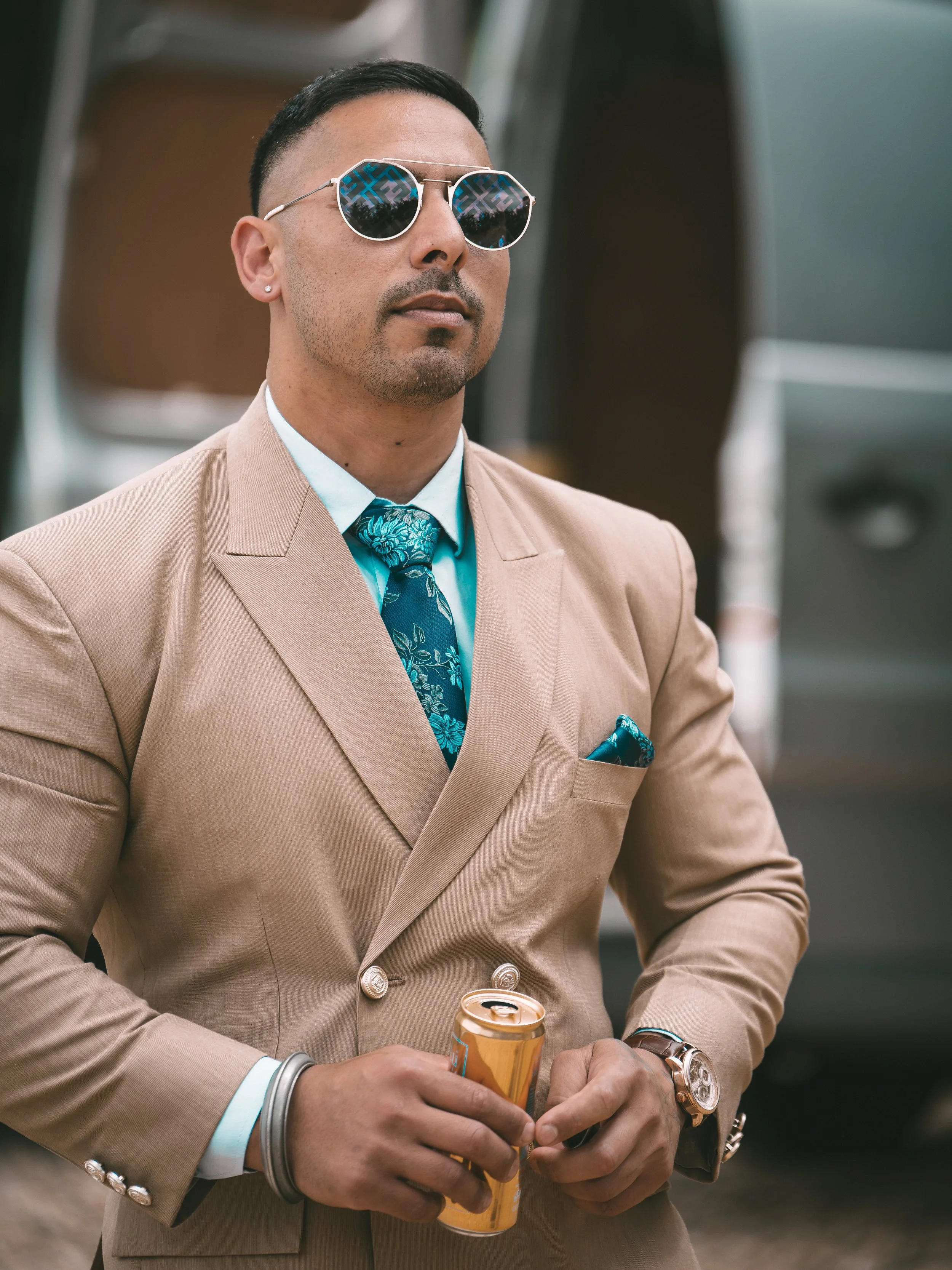 A man wearing a beige suit, patterned teal tie, and sunglasses holds a can of beer. He has earrings, a watch, and several bracelets.