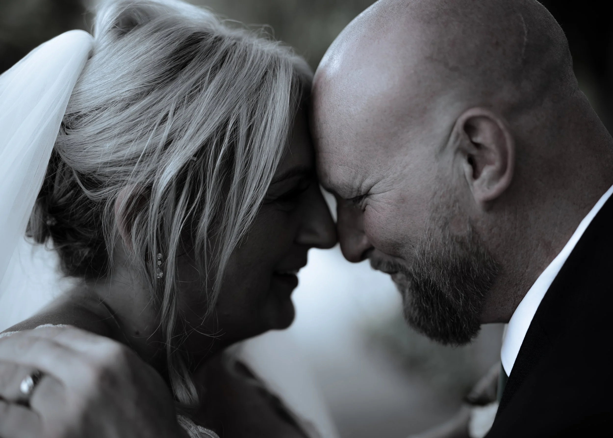 Close-up of a couple, a woman with long grey hair and a man with a shaved head, touching foreheads and eyes closed, in a tender moment.