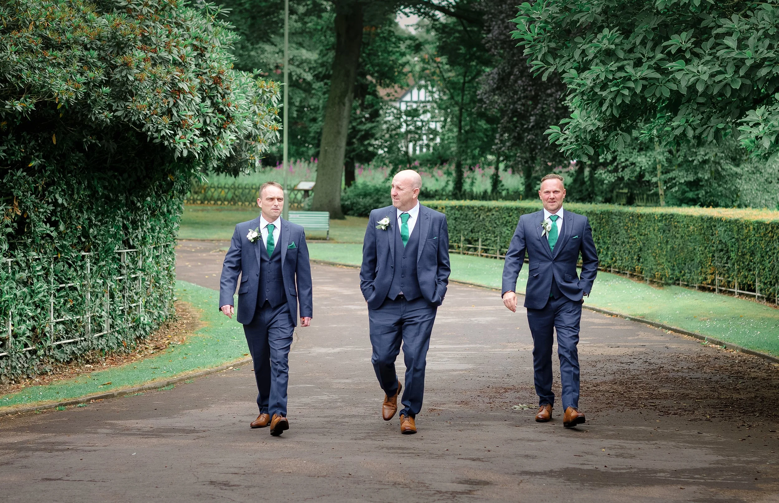 Three men dressed in navy blue suits with white shirts, green ties, and boutonnières walk through a park with lush green trees and hedge bushes, during daytime.
