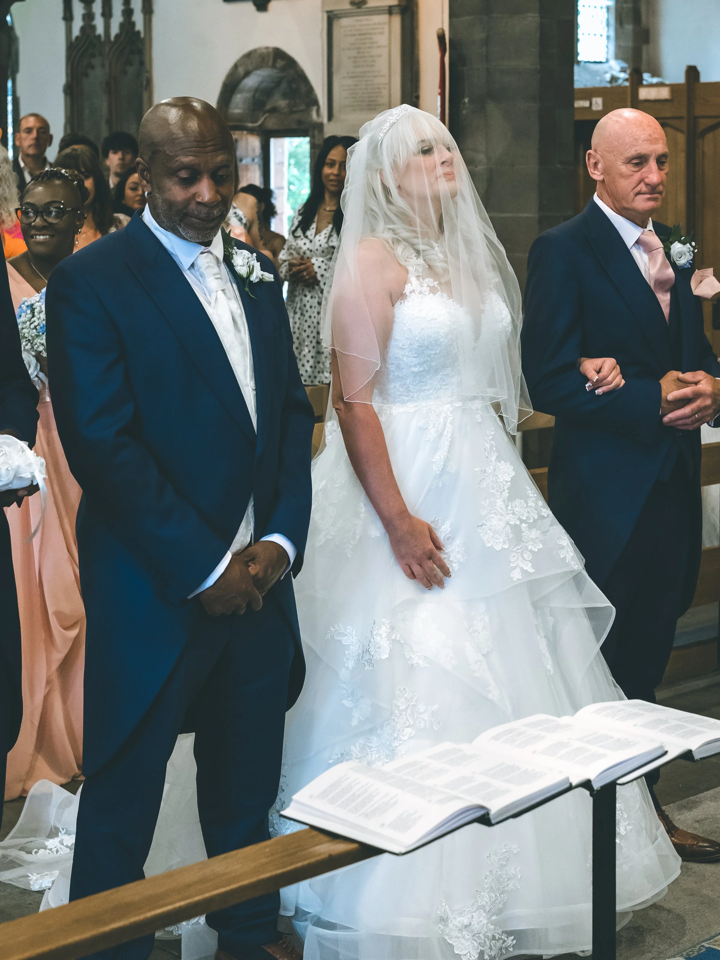 A group of people, including a bride in a white wedding dress and veil, and two men in suits, are participating in a wedding ceremony inside a church. They are standing with eyes closed, appearing to read or recite their vows from an open book on a s