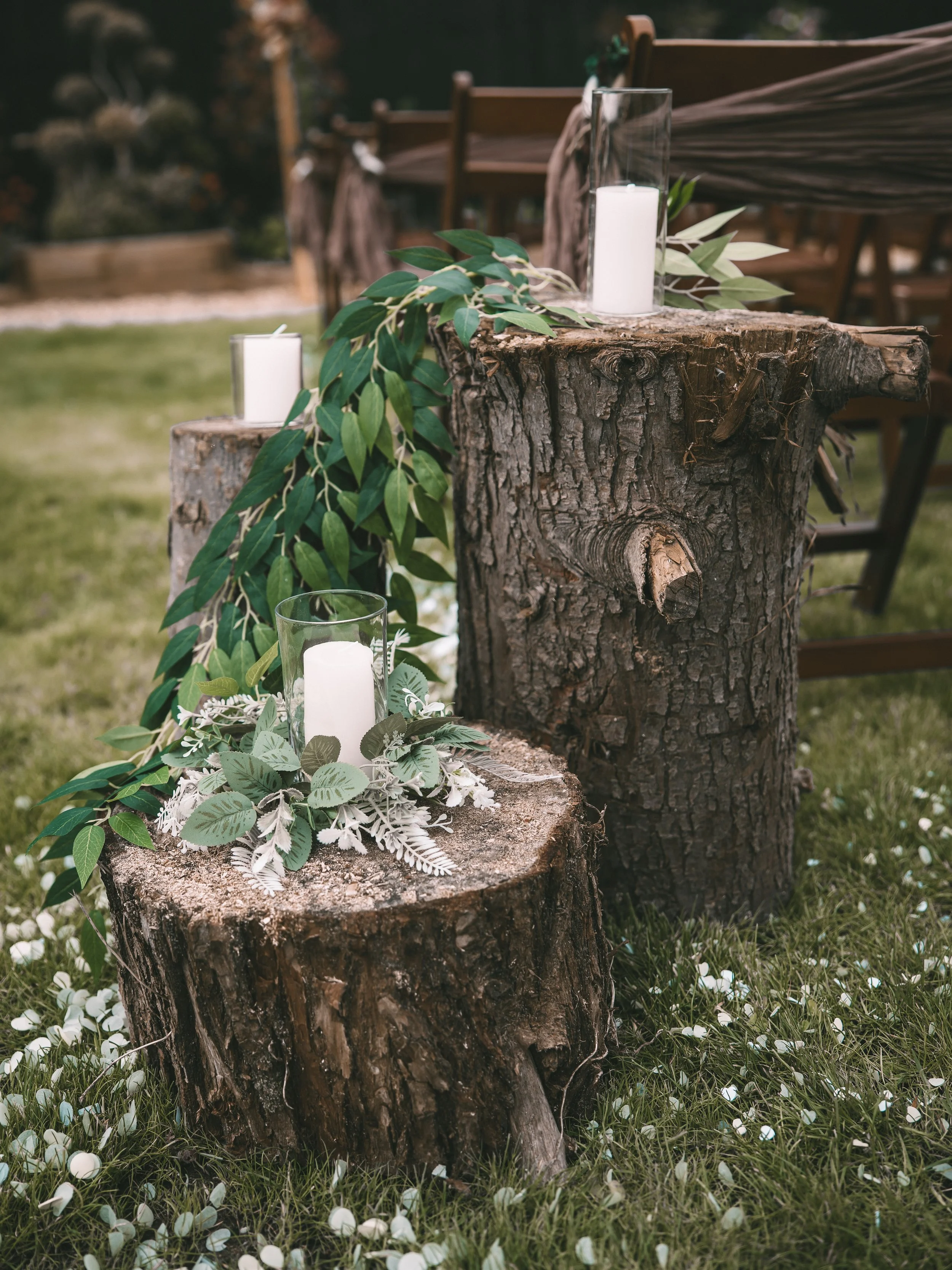 A decorative outdoor setup featuring tree stumps with candles in glass holders and greenery on top, surrounded by white flower petals on the grass.