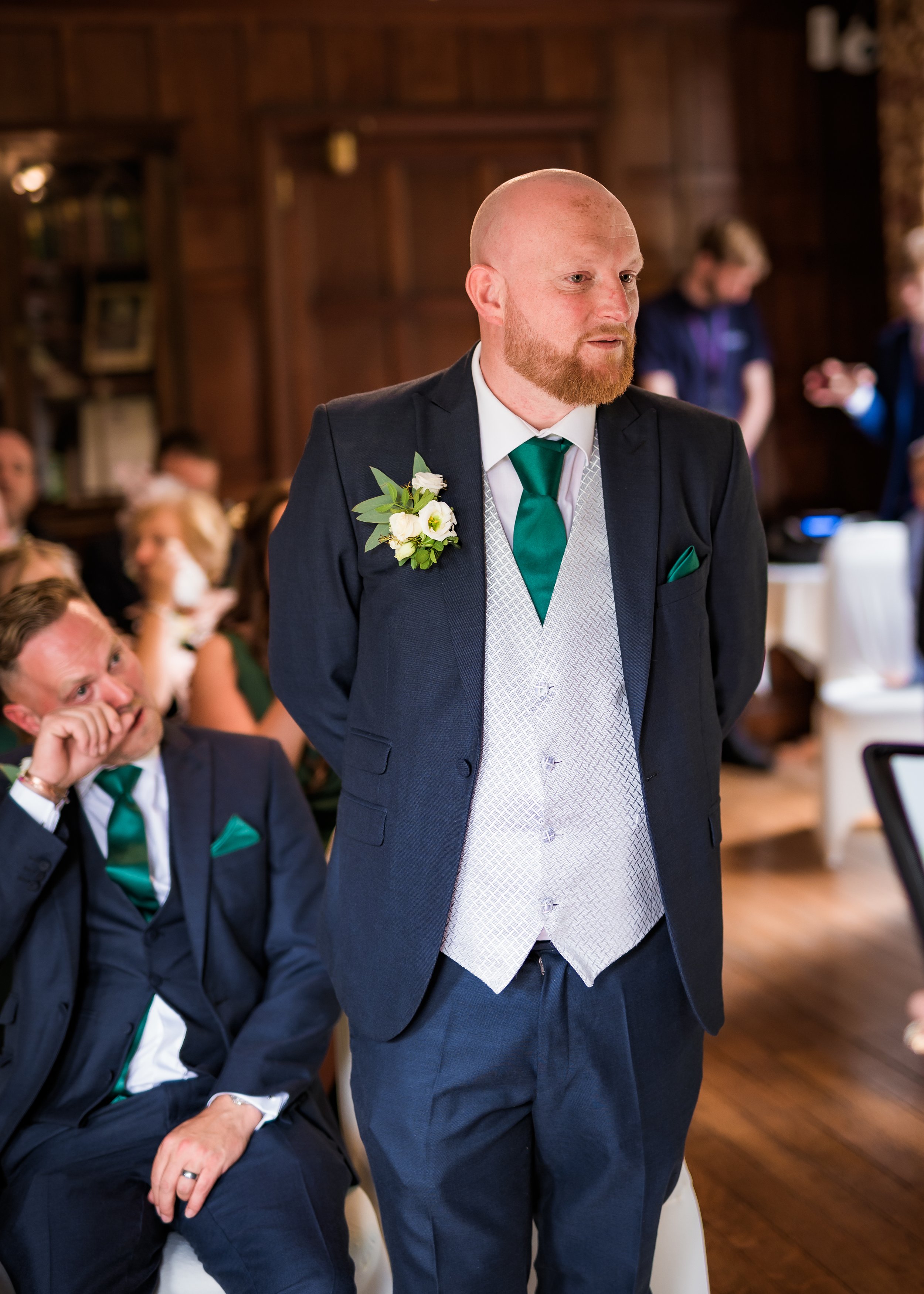 A man with a bald head and beard, dressed in a dark suit, white patterned vest, white shirt, and green tie, standing in a room with wood-paneled walls during a formal event. He wears a boutonniere with white flowers and green leaves on his left lapel