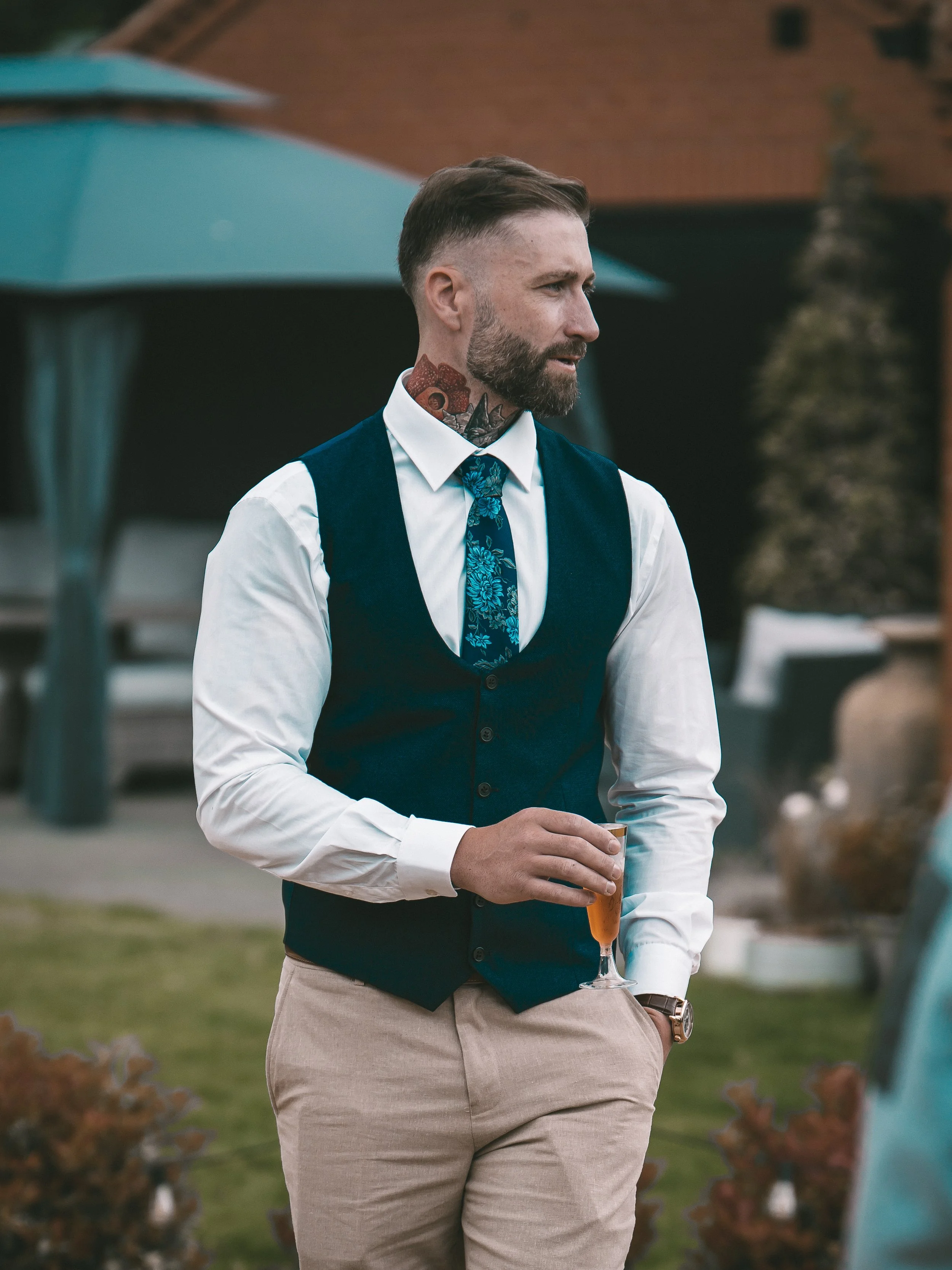 A man with a beard and a tattoo on his neck, wearing a white shirt, blue vest, and floral tie, is holding a glass of beer during an outdoor event.