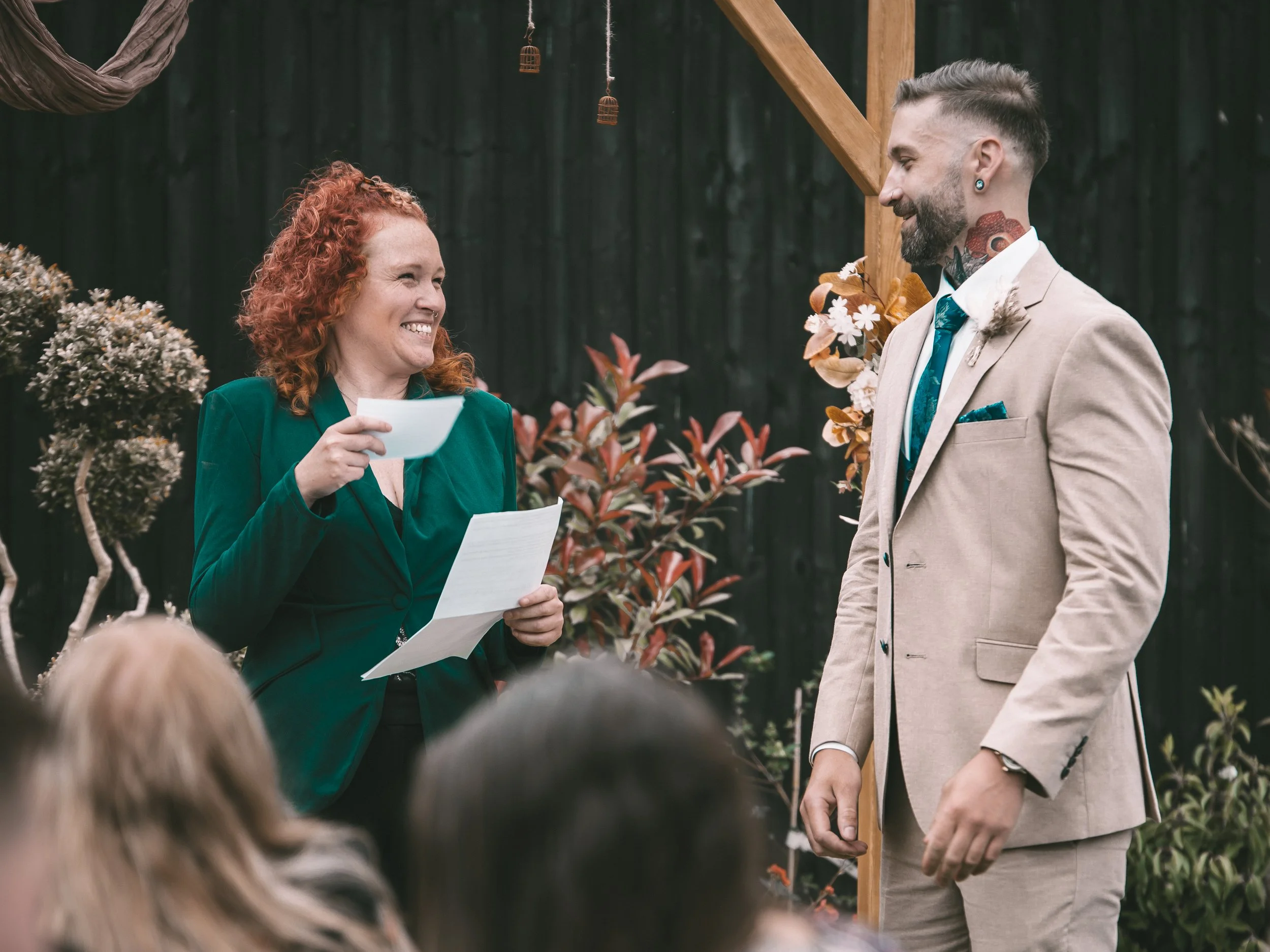 A couple at a wedding ceremony, the woman with curly red hair in a green jacket holding a piece of paper, and the man with a beard and tattoo on his neck wearing a beige suit, exchanging vows outdoors in front of a dark fence and greenery.