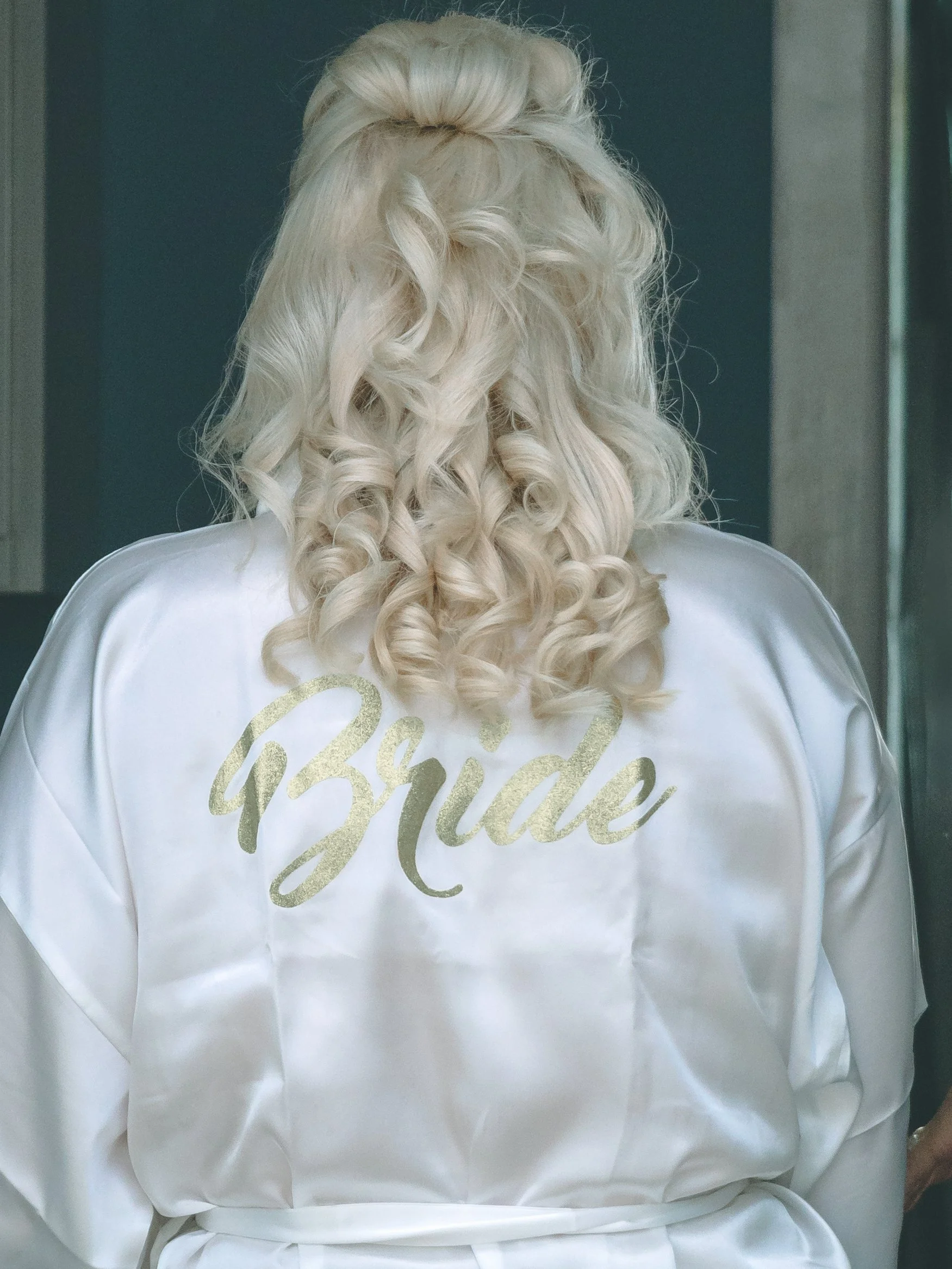 Back of a woman with platinum blonde, curly hair styled in an updo, wearing a white satin robe with gold lettering that says 'Bridal'.