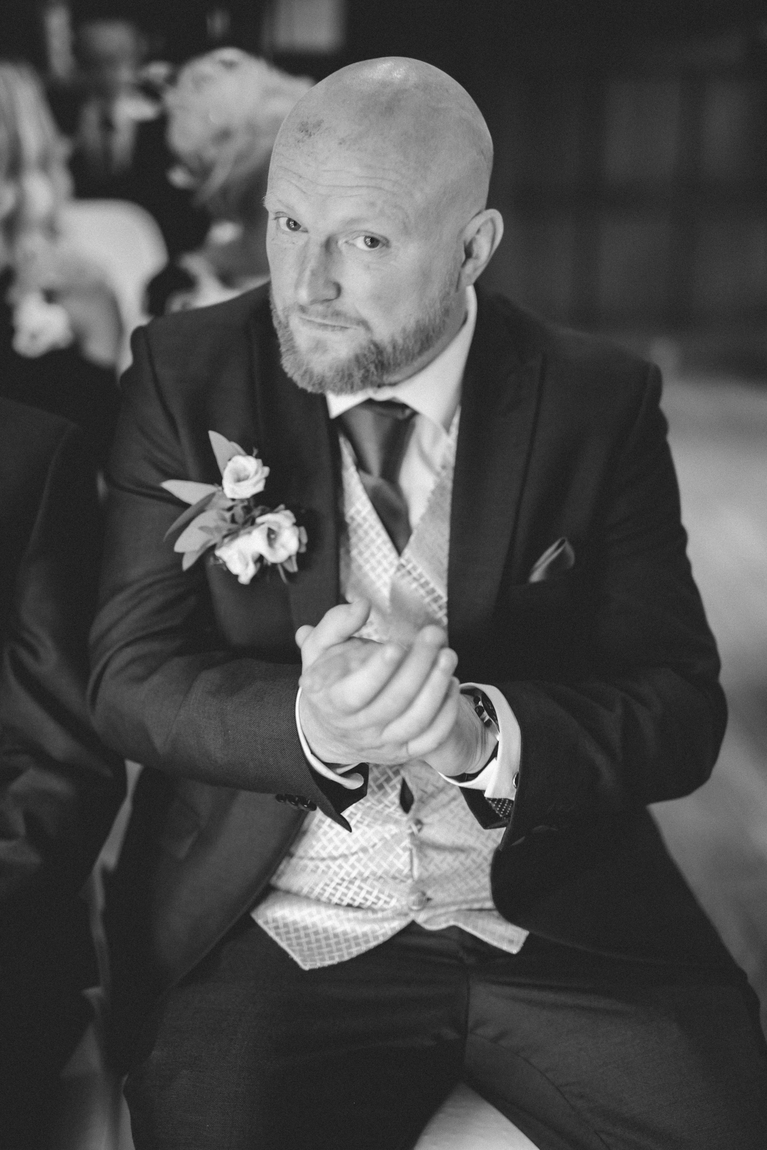 Black and white photo of a bald man with a beard, wearing a suit, tie, and a boutonniere, sitting at a table with his hands clasped, with people blurred in the background.