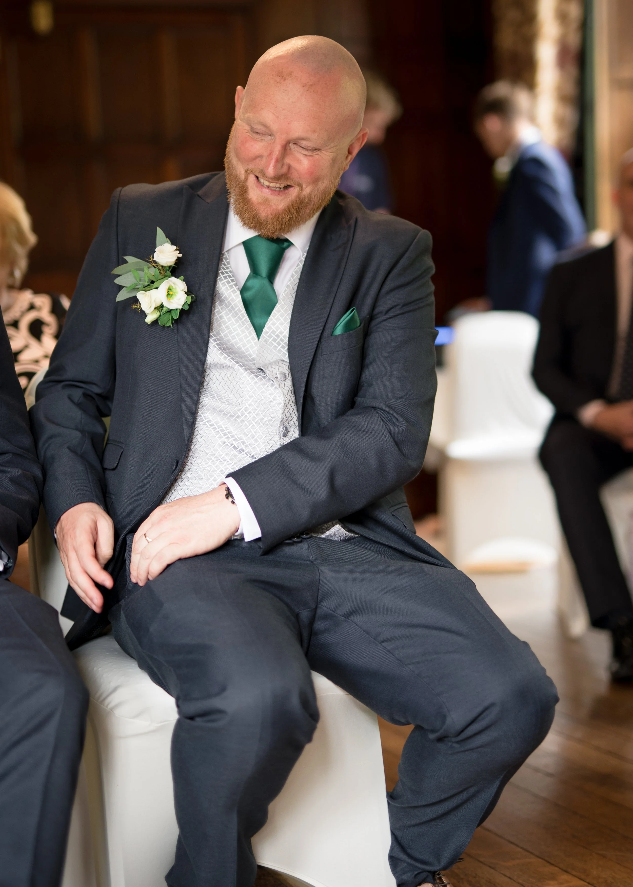 A happy man in a suit with a green tie and boutonniere sitting in a chair at a formal event, surrounded by other people.
