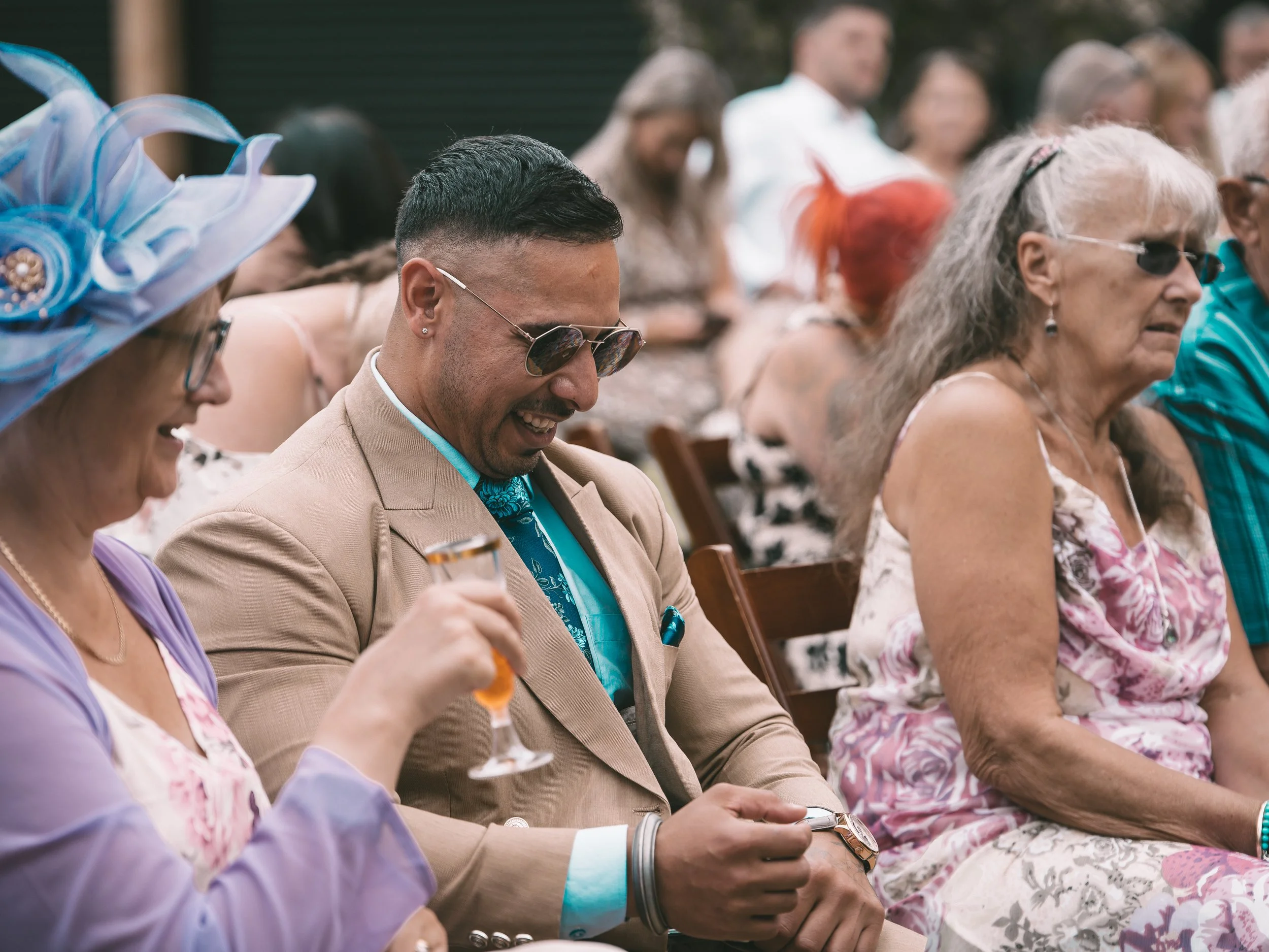 People sitting in rows at an outdoor event, smiling and holding drinks, dressed in colorful, summery clothing.