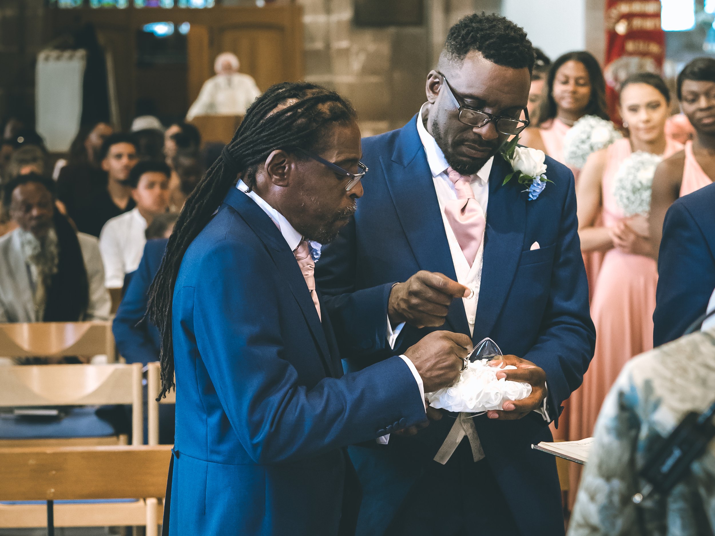 Two men in blue suits, pink ties, and white shirts stand closely together during a wedding ceremony inside a church. They are looking down and holding a small box with white flowers. People in formal attire are seated in the background.