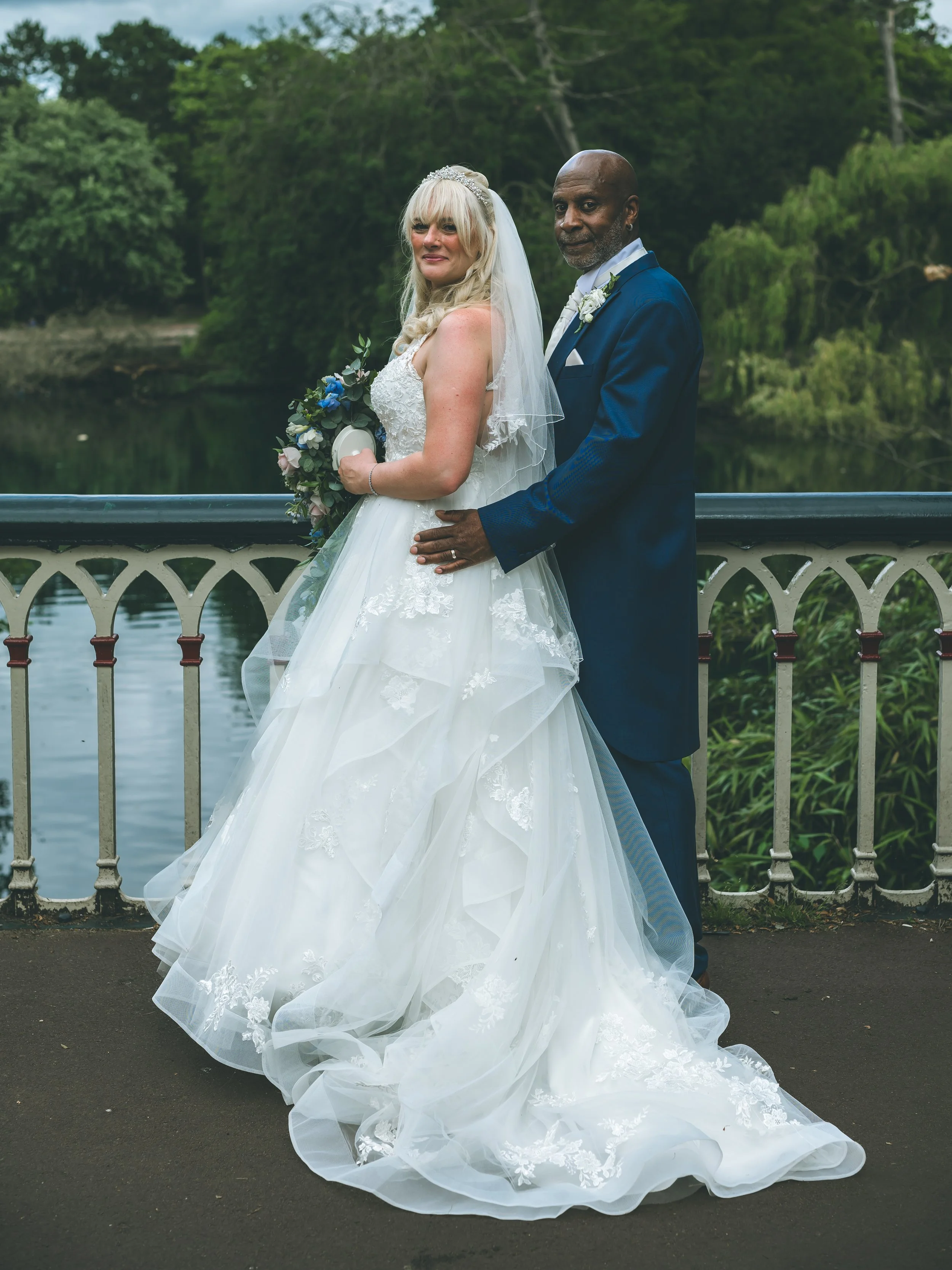 A bride in a white wedding gown holding a bouquet, standing next to her groom in a blue suit with a white boutonniere, on a bridge by a body of water with greenery in the background.