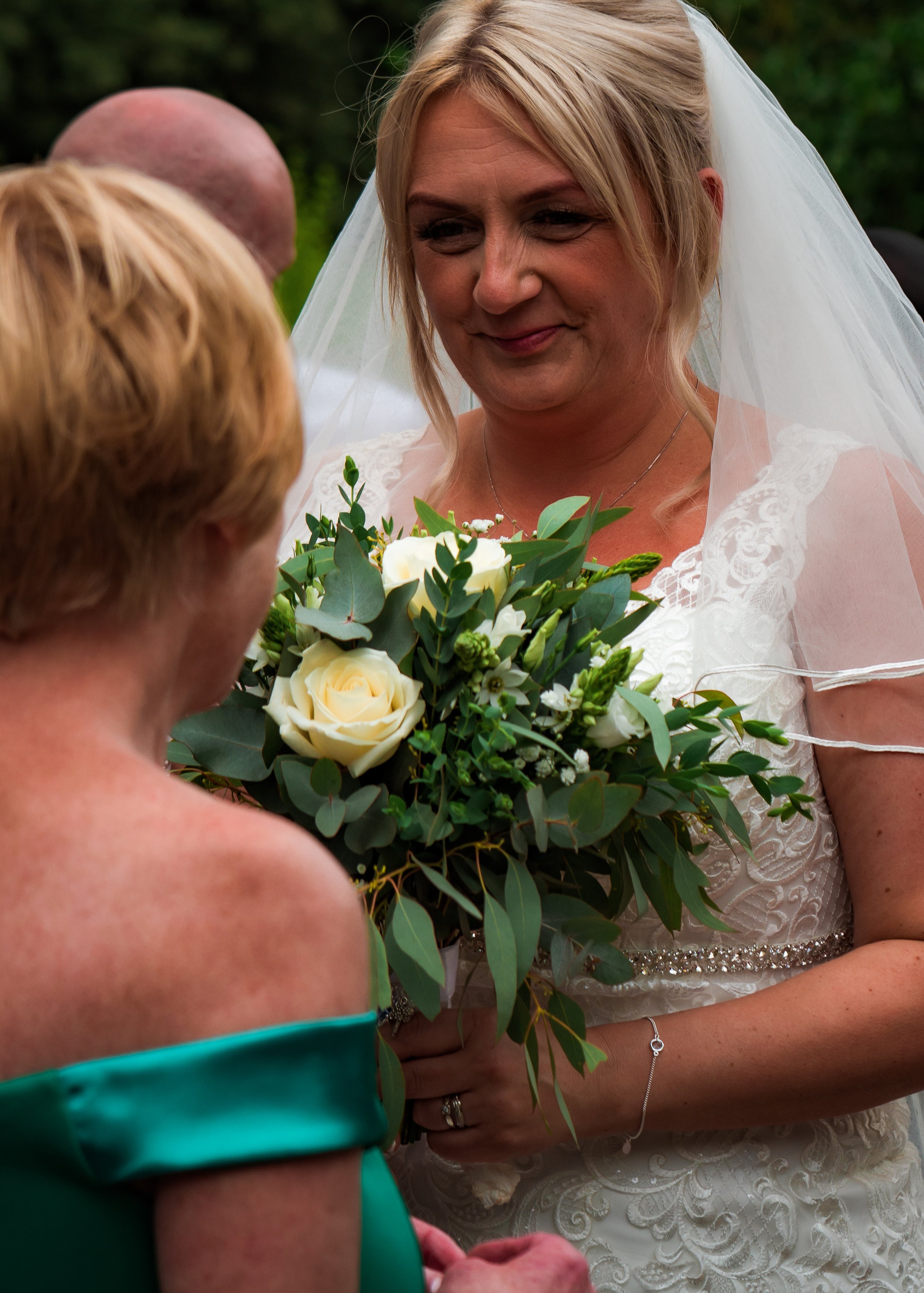 A bride in a white lace wedding dress with a veil is holding a bouquet of white roses and greenery. She is smiling softly and looking down at someone in front of her, outdoors with a blurred green background.