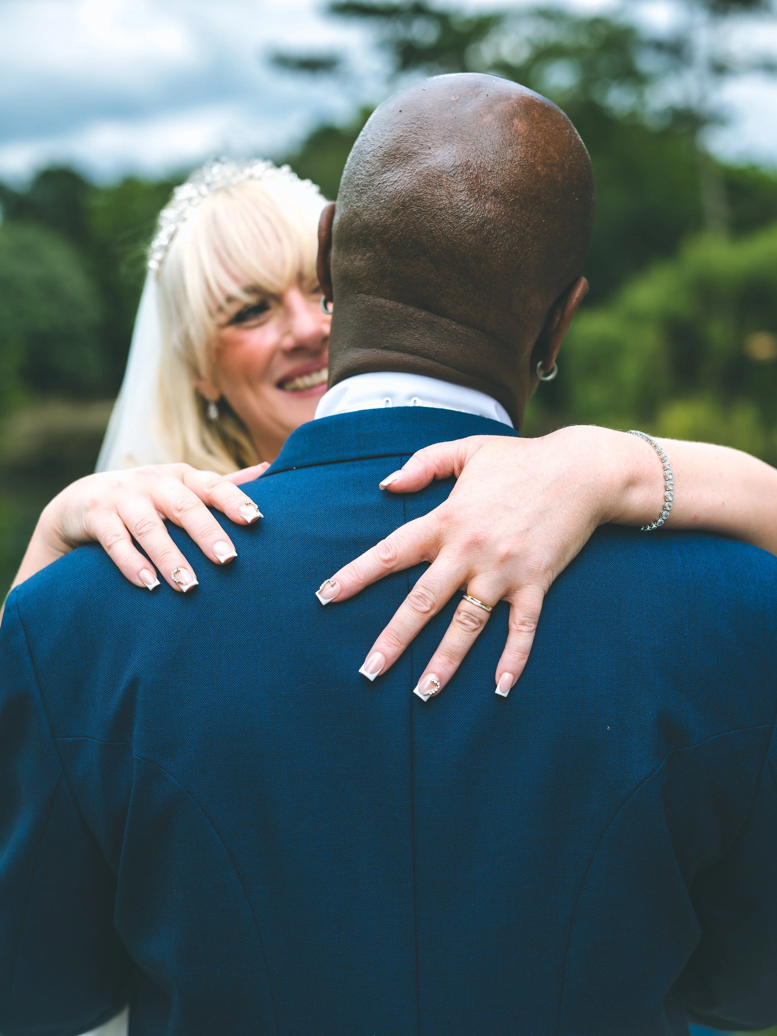 A bride and groom embrace outdoors, with the bride smiling and wearing a floral headpiece, while the groom wears a dark blue suit; focus on their upper bodies and arms.
