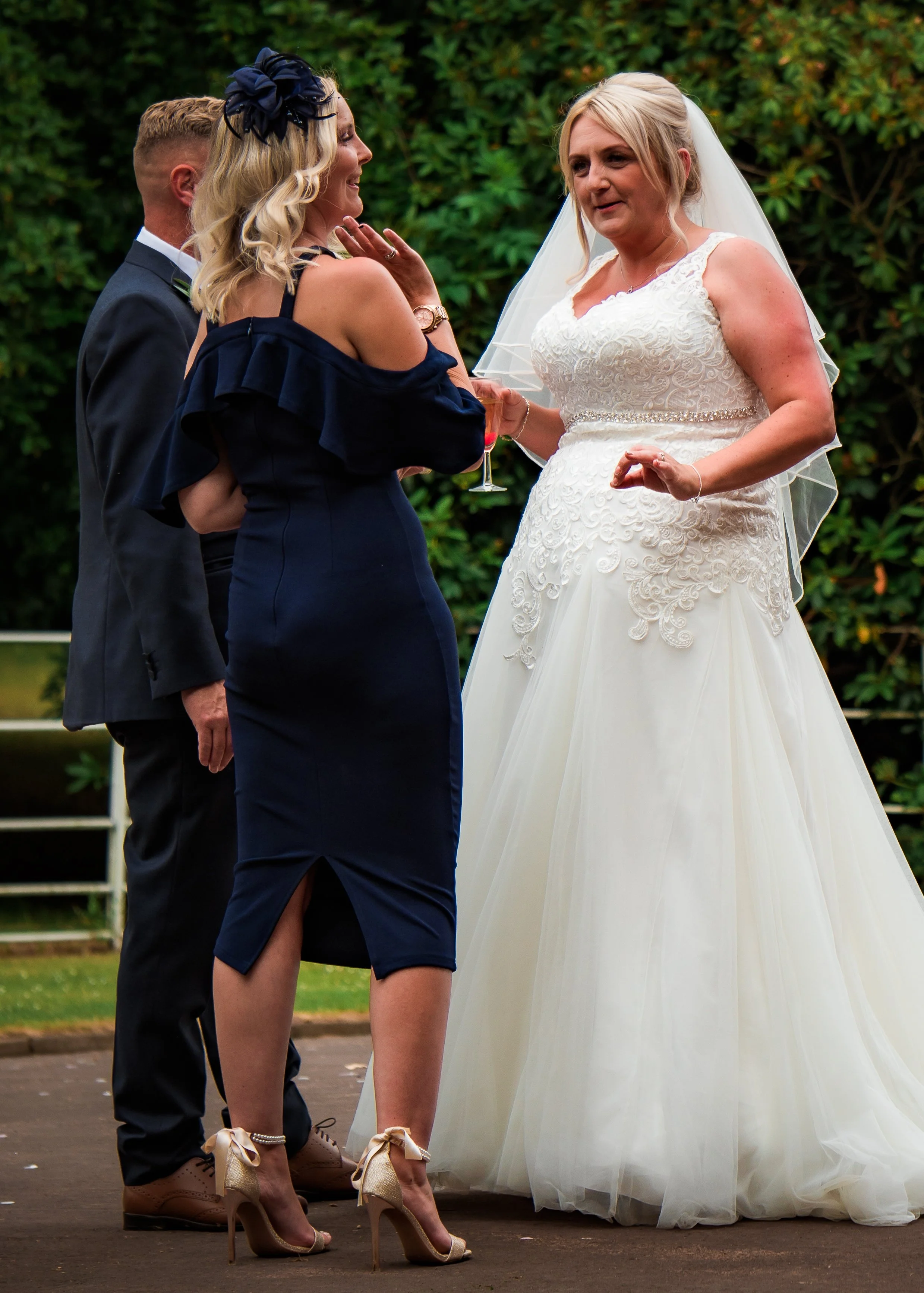 A bride in a white wedding gown standing outdoors and talking with a woman in a navy blue dress with off-shoulder ruffles. Two men are partially visible behind them, one in a suit. The background features green bushes and a fenced area.