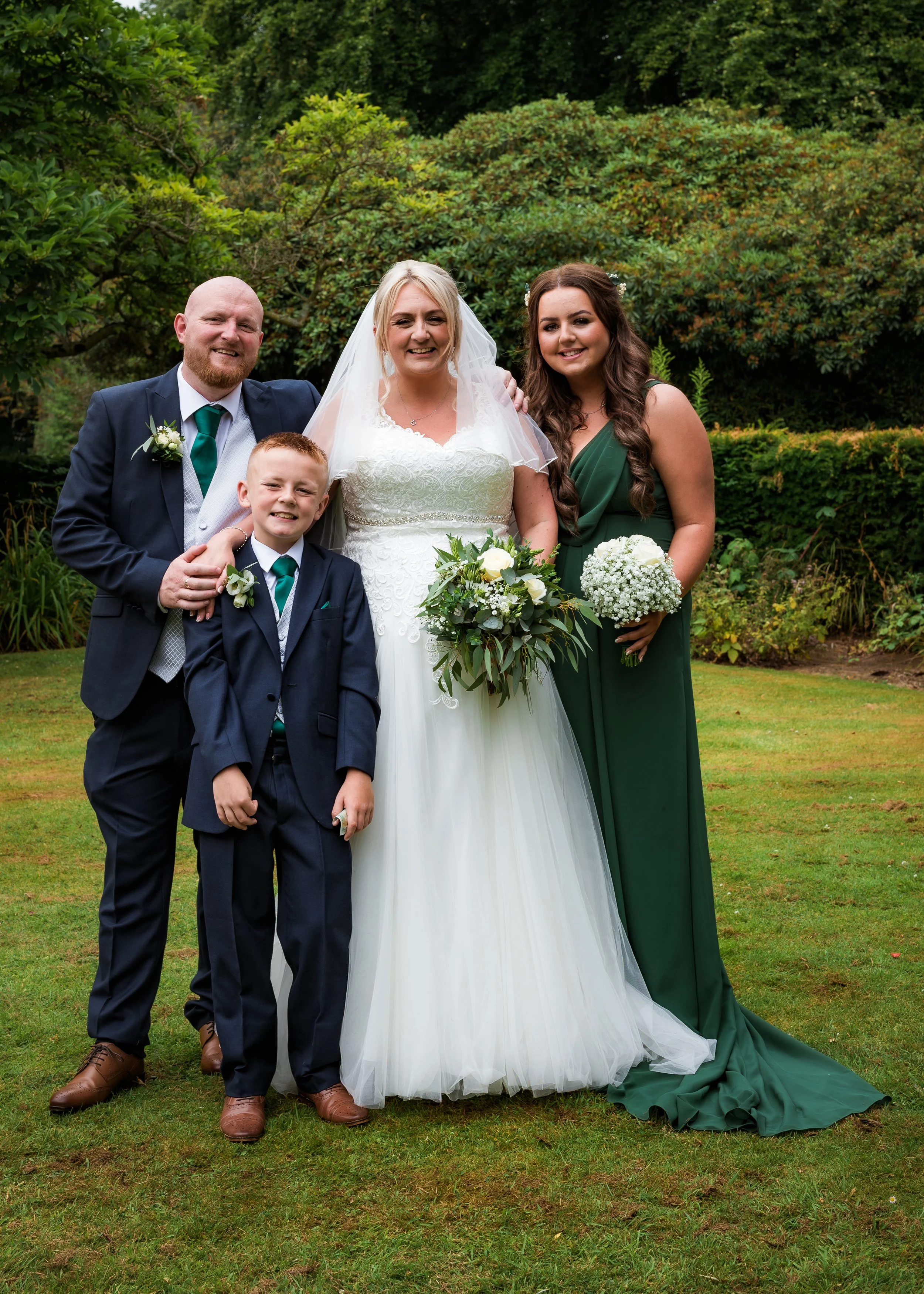 A smiling bride in a white wedding dress with a veil, standing with a groom, a woman in a green dress, and a young boy, outdoors on a lawn with green bushes and trees in the background.
