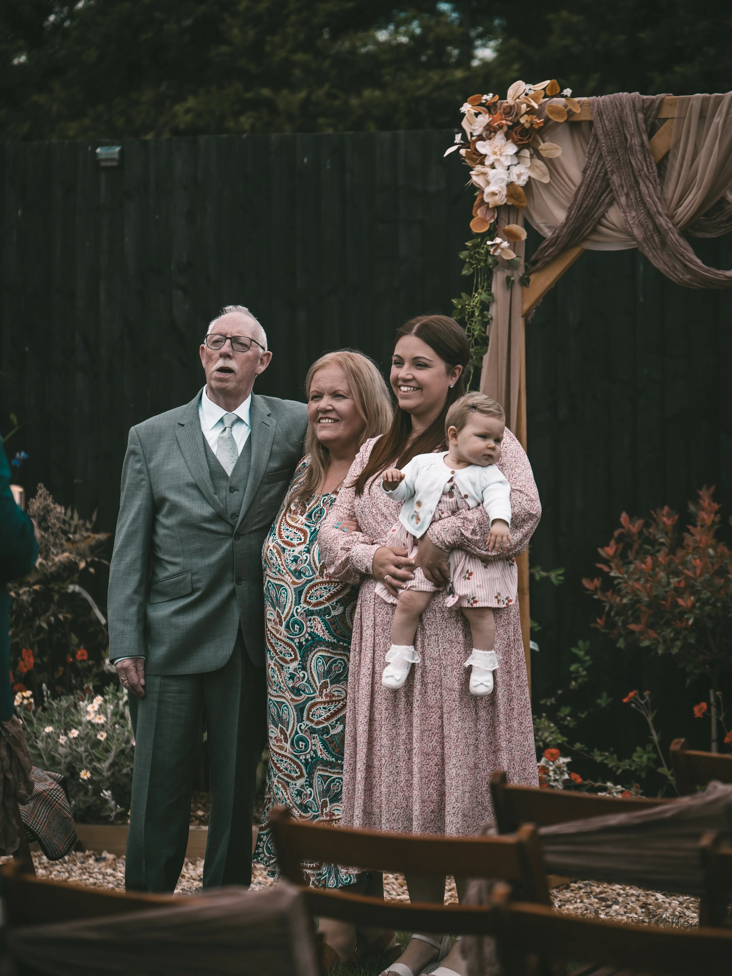 Group photo of four people, including an elderly man, a middle-aged woman, a young woman, and a baby girl, posing outdoors in front of a decorated wooden arch during a celebration or event.